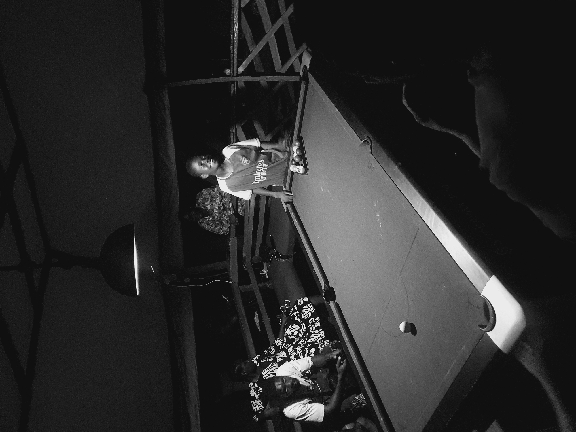 A monochrome, high-angle shot shows a pool table indoors, presumably in Lokoja, Nigeria, given the prompt. A young man in a jersey stands on the left side of the table, smiling. Balls are racked on the table's upper right side. Below him, two people are seated, one in a patterned shirt, the other in a white shirt. A single ball is near a pocket on the table. A lamp hangs to the left, while parts of a wooden structure are visible above the pool table. The surrounding area is dark, with faint outlines of people on the right.