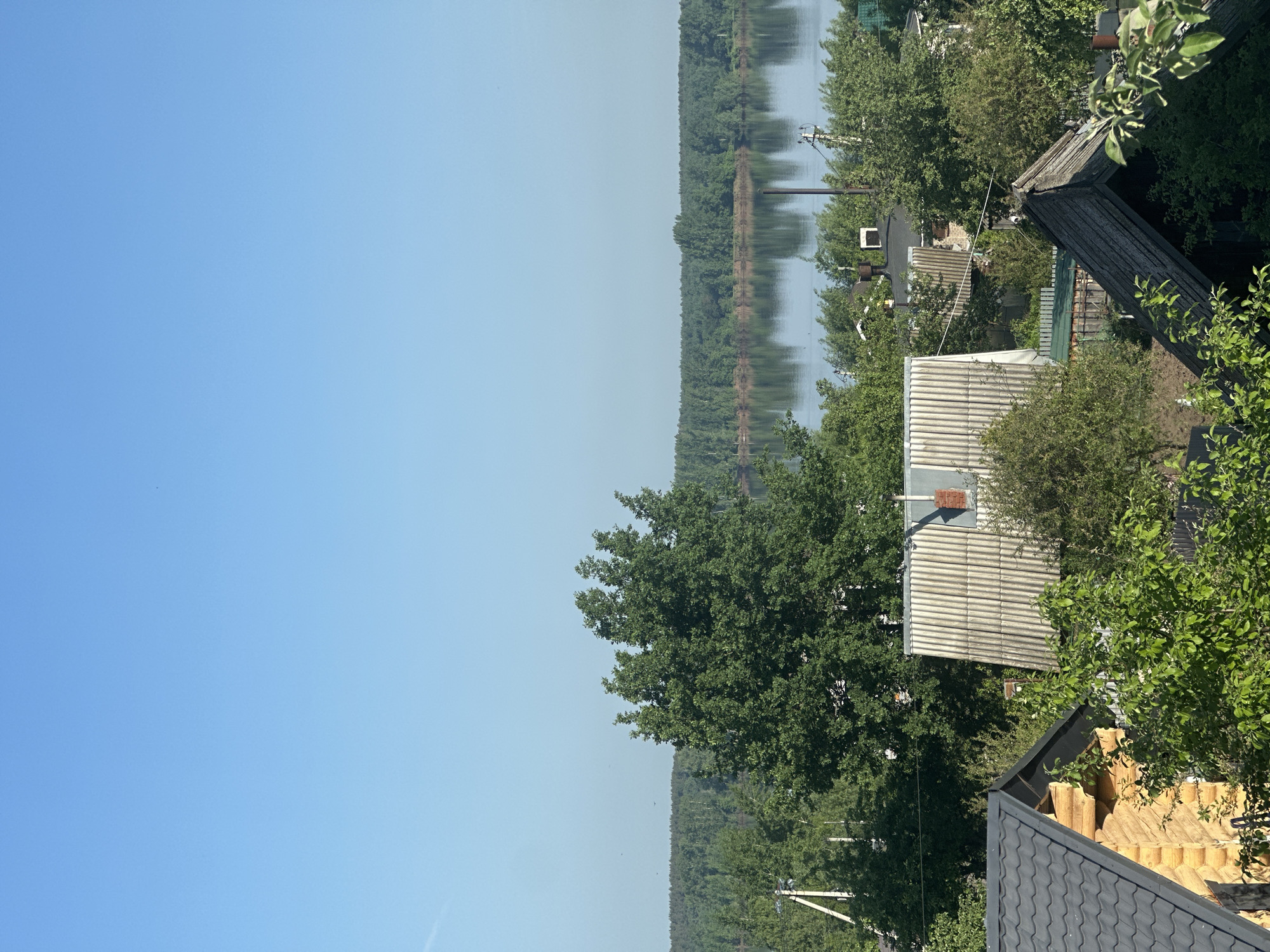 The image shows a view of the Kama River in Chaikovsky, Russia. The scene is captured from an elevated vantage point, possibly a rooftop or upper story of a building. The river is visible in the background, bordered by a dense line of trees along the opposite bank, creating a natural boundary.
In the foreground, there are several residential houses with varied roof types. One roof is made of gray corrugated metal, while another is a lighter color, likely made of slate or a similar material. Green foliage, including trees and bushes, is interspersed around the buildings, suggesting a blend of residential and natural environments. Utility poles and wires are visible, indicating the presence of infrastructure in the residential area.
The sky is clear blue, suggesting a sunny day. There are no visible people in the frame. The overall scene is a landscape featuring a river, residential buildings, and natural elements.