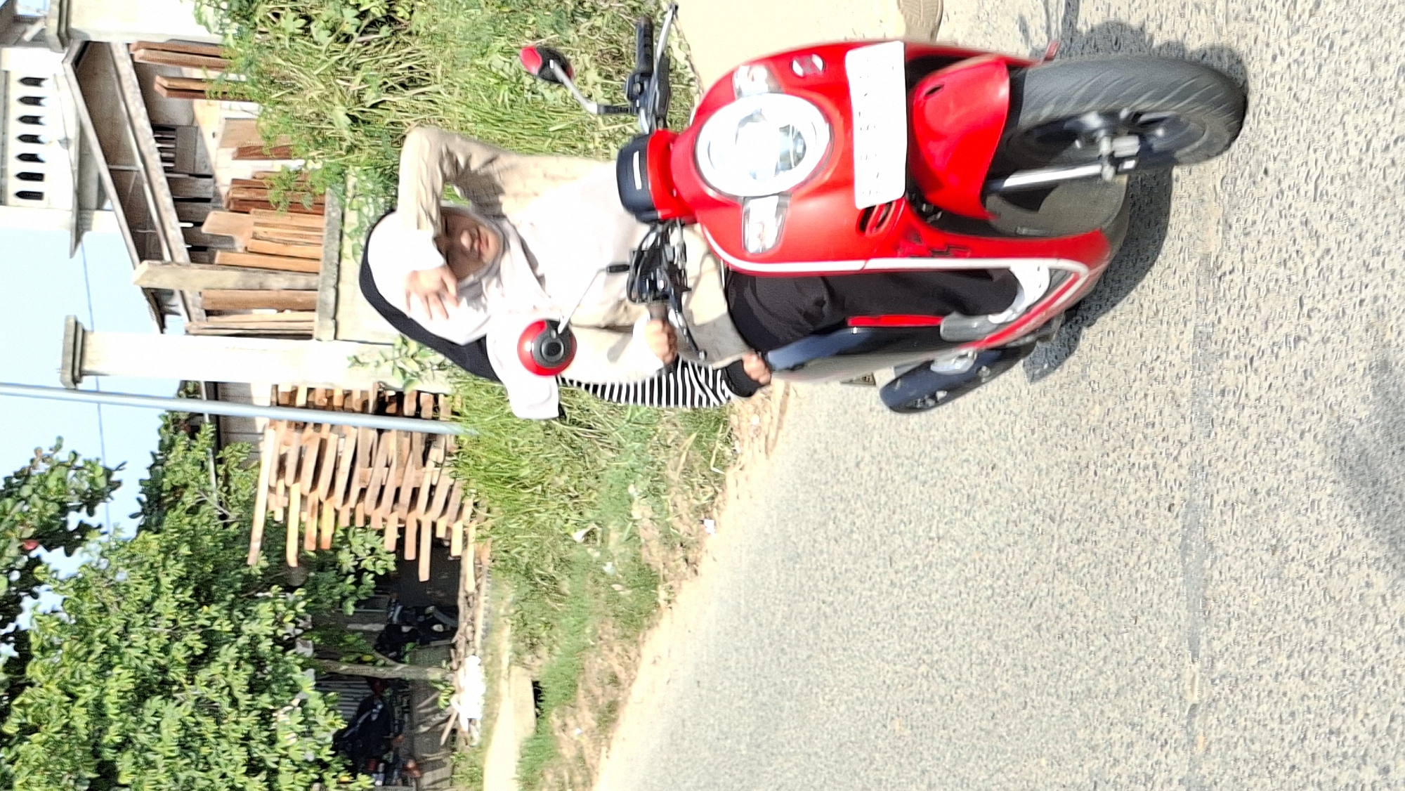 A young woman wearing a light-colored hijab and a tan jacket is riding a red scooter on a paved road. She is shielding her eyes with her right hand from the sun. She is wearing black pants and a striped shirt underneath the jacket. The scooter has a white license plate that is partially obscured. The road is flanked by a grassy area with some scattered debris and a pile of wooden planks. In the background, there are trees, a building that appears to be a mosque with a distinct architectural style, and more wooden planks stacked vertically. The sky is clear and blue. This scene is likely located in the city of Lhokseumawe, Indonesia, given the architectural style of the building and the overall environment.