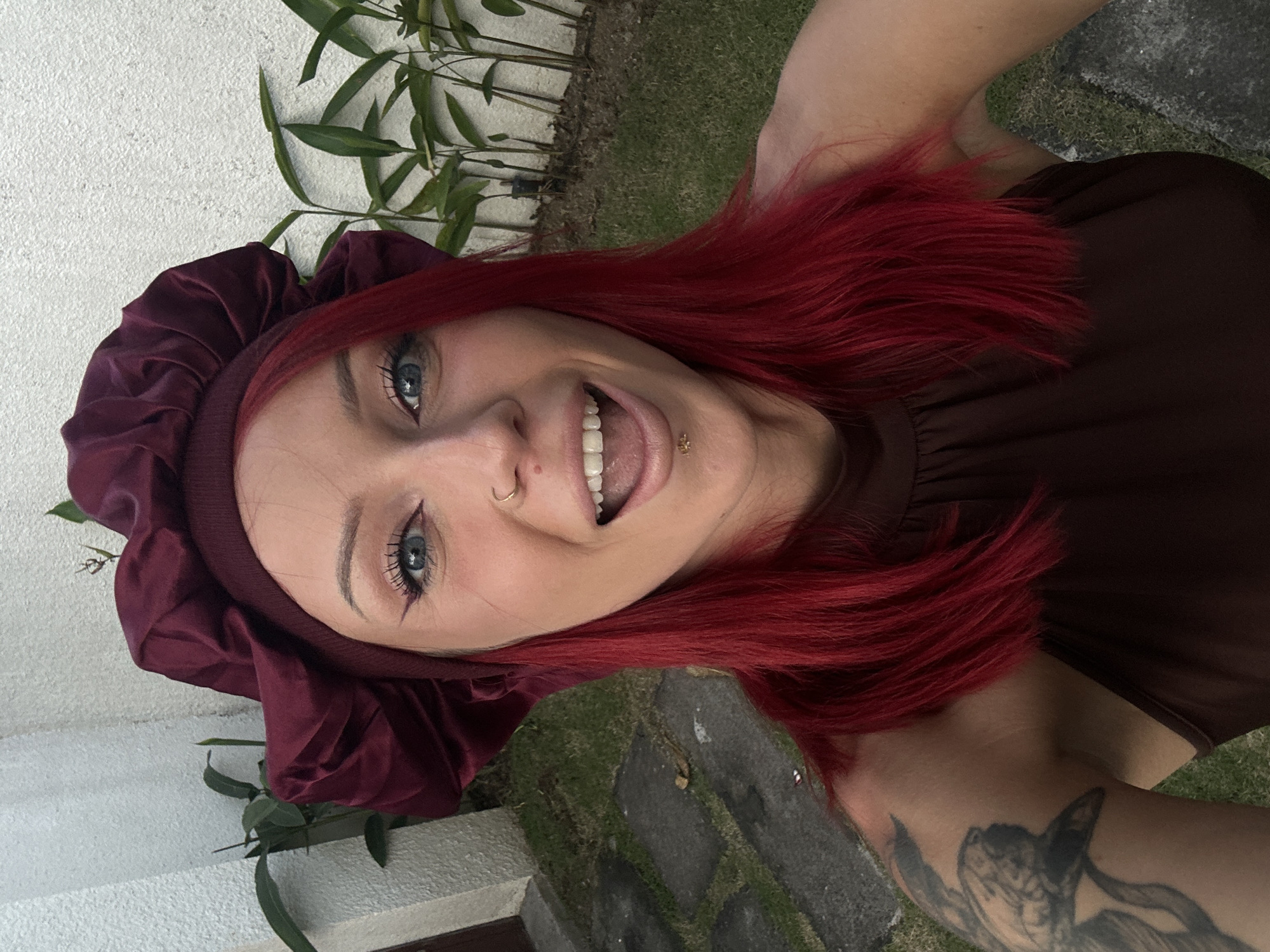 A woman with bright red hair and blue eyes poses outside in North Kuta, Indonesia. She is wearing a dark purple bonnet, a brown top, and has a tattoo on her arm. She has a nose ring and a lip piercing and is sticking her tongue out. A white wall and green plants are in the background, along with grass and a stone path.
