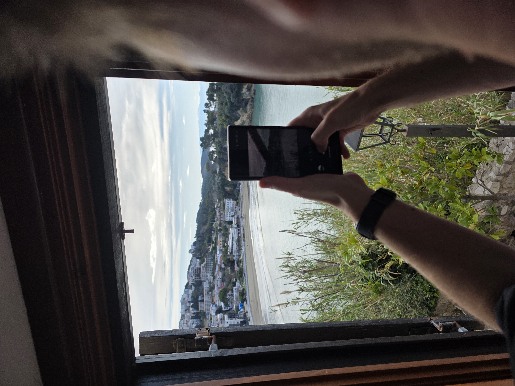 The scene captured is a photograph taken through what appears to be an open window, partially obscured by the photographer's body. Outside the window is a view of a coastline city, presumably the city of Ulcinj - Ulqin, Montenegro.
A person is holding a smartphone, seemingly taking a picture of the coastal view. The smartphone screen is dark. The person is wearing a black smart watch on their left wrist. Their arms and part of their torso are visible, framing the scene.
The coastal view includes buildings clustered along the shoreline and up the hillside. There is a sandy beach and the ocean visible, with a headland or peninsula extending into the sea, covered with greenery. The sky is overcast, with light clouds. Green foliage and a wrought iron railing are visible just outside the window. The window frame is made of dark brown wood.