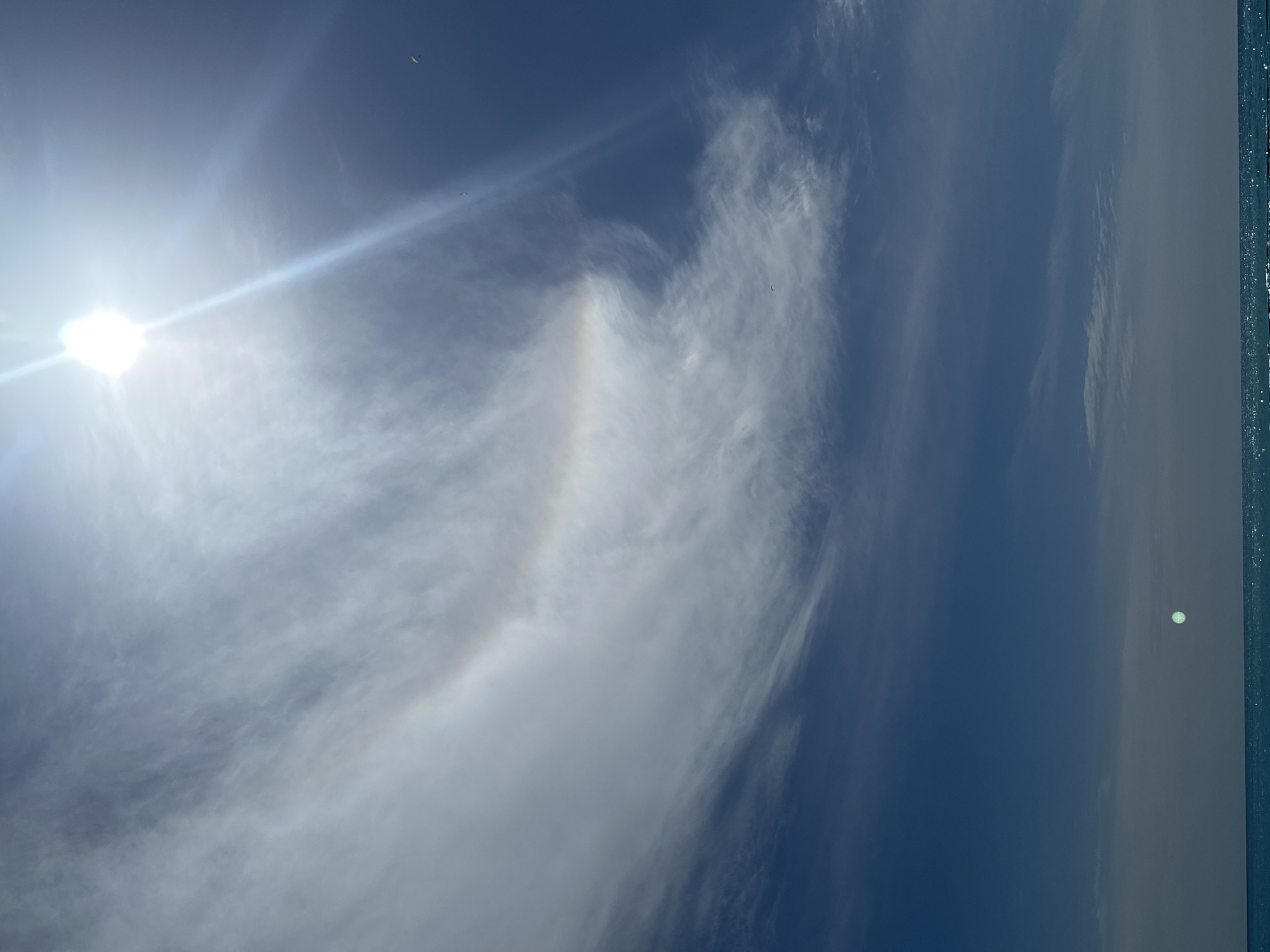 The image depicts a daytime sky scene in Fethiye, Turkey. A bright sun is visible in the upper left, creating a lens flare effect. A halo effect, possibly a 22° halo, is partially visible in the surrounding cirrus clouds. Several objects, possibly paragliders, are scattered throughout the scene. The edge of a building or structure with reflective details is visible on the right.