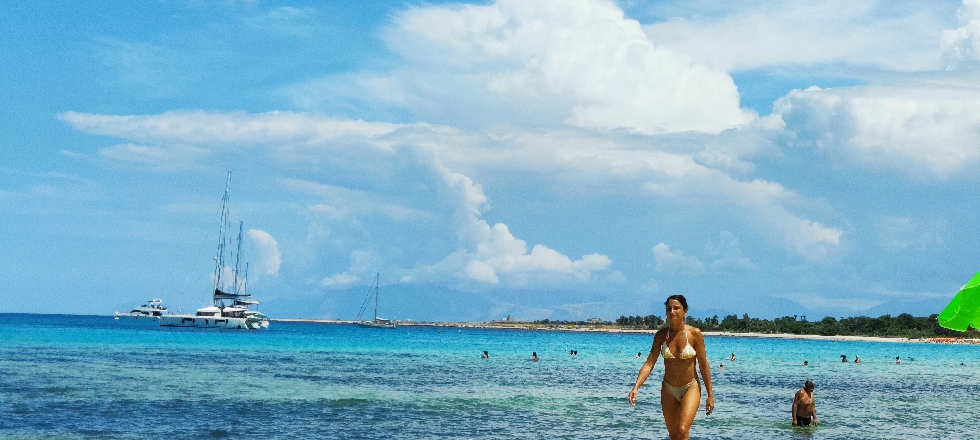 This is a wide panoramic shot of a beach in San Vito Lo Capo, Italy. The foreground shows shallow, clear turquoise water with some ripples. A woman in a gold bikini is walking towards the right of the frame through the water, with her head turned slightly to the right. Further out in the water, several other people are visible, some swimming and wading. In the mid-ground, two sailboats are anchored, one a large white catamaran with multiple masts and another a smaller white sailboat to its left. The background features a coastline with green trees and some buildings. The sky is a vibrant blue with large, puffy white clouds. The scene is set on a sunny day.