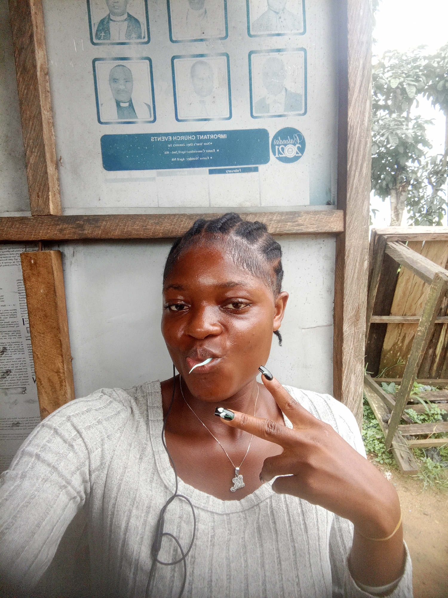 The image shows a young, dark-skinned woman taking a selfie outdoors. She is wearing a light-colored, long-sleeved shirt and a silver necklace with a pendant. She has her hair styled in braids and is holding a white object in her mouth, possibly a toothpick. Her nails are painted with a black and white design. She is making a peace sign with her right hand.
Behind her is a bulletin board with photographs of men in suits, and a sign with text in English and possibly another language. The text includes "IMPORTANT CHURCH EVENTS" and a logo that reads "ISOS 2021." The bulletin board is framed with rough-hewn wood.
To the right of the woman is a wooden structure that appears to be a broken or makeshift staircase, surrounded by green grass and foliage. The image was taken in Mbribit, Nigeria.