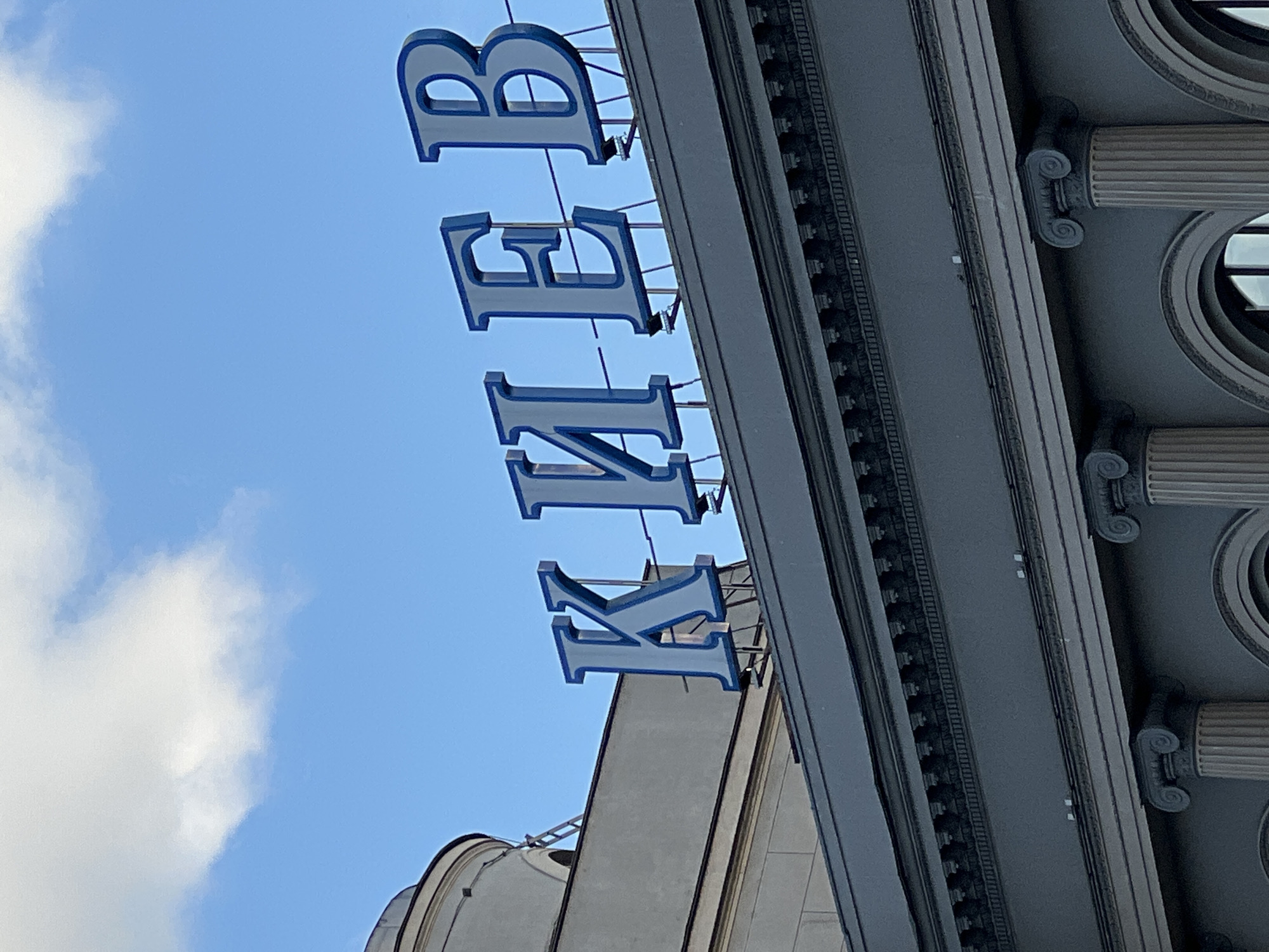 The image shows a close-up, angled shot of a building's facade with the word "КИЕВ" (KIEV) displayed in large blue and white letters across the top. The letters are mounted on the edge of the roof and are illuminated. The building is a dark, classical-style structure with fluted columns and decorative molding visible below the letters. The background is a clear blue sky with some white clouds. The scene is observed from a low angle, looking upwards at the signage. There are no people visible in the image. The architecture and the signage suggest this is likely a significant building, possibly a train station or a public institution. This specific signage and architectural style are characteristic of buildings in Moscow, Russia.