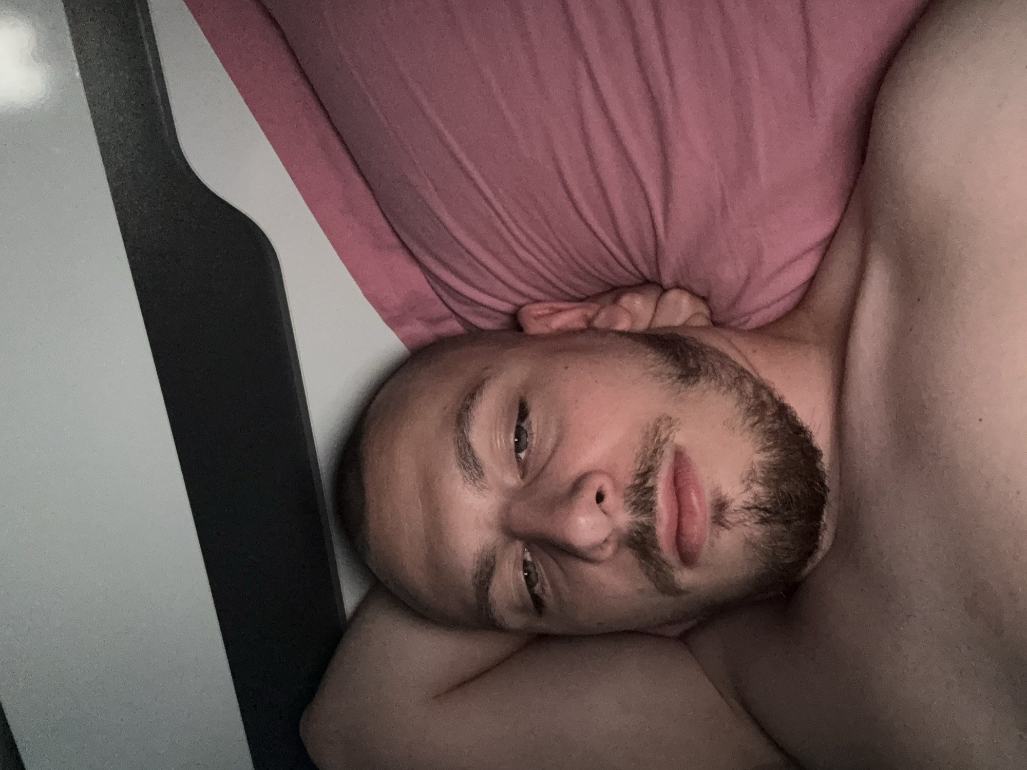 This image depicts an indoor scene in Stara Zagora, Bulgaria. A male individual, appearing to be a young adult, is shown from the chest up, lying down. He has a closely shaven head and a short, neatly trimmed beard and mustache. His eyes are partially open, with a direct but relaxed gaze towards the viewer. His right arm is bent, with his hand positioned behind his head or pillow on the left side of the frame. He appears shirtless, exposing his bare chest and shoulder. His head rests on a pink or mauve-colored pillow, which exhibits various folds and wrinkles in its fabric. Above the subject's head, a white surface is visible, horizontally bisected by a dark grey or black strip, consistent with a headboard or wall-mounted furniture. The scene is dimly lit with a soft, warm light, indicating an indoor, low-light environment.