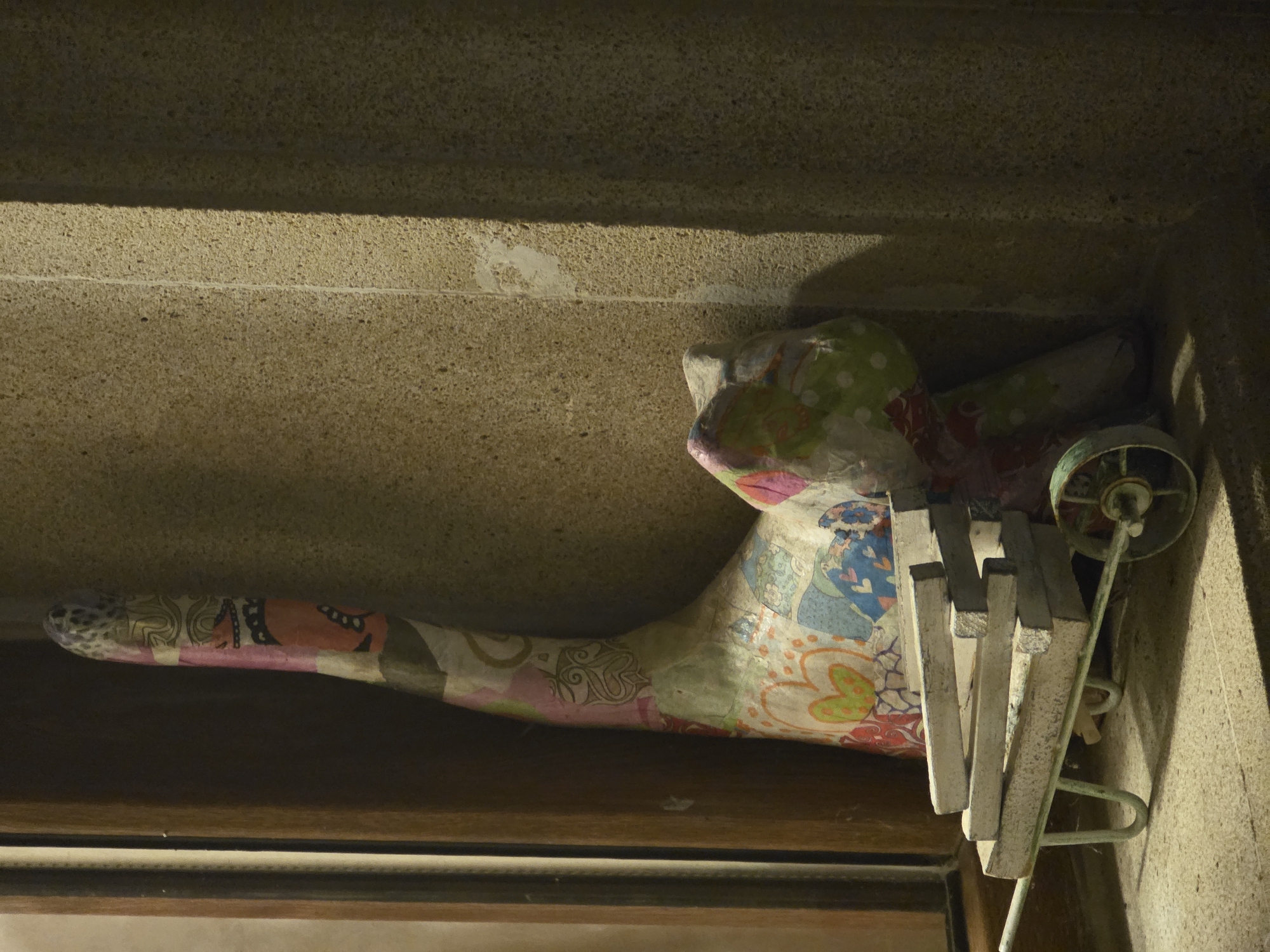 The image shows a colorful, patchwork doll-like sculpture leaning against a textured stone wall. The sculpture has a long, curved neck adorned with various patterns, including floral motifs and abstract designs. A small, weathered green wheelbarrow, stacked with five rectangular wooden planks, is positioned in front of the sculpture. The wheelbarrow's handles extend to the left, and one wheel is visible on the right. The environment appears to be outdoors, with a wooden doorframe visible on the left. The lighting suggests it might be dusk or early morning, casting shadows and highlighting the textures of the stone. The scene is located in Buzovna, Azerbaijan.