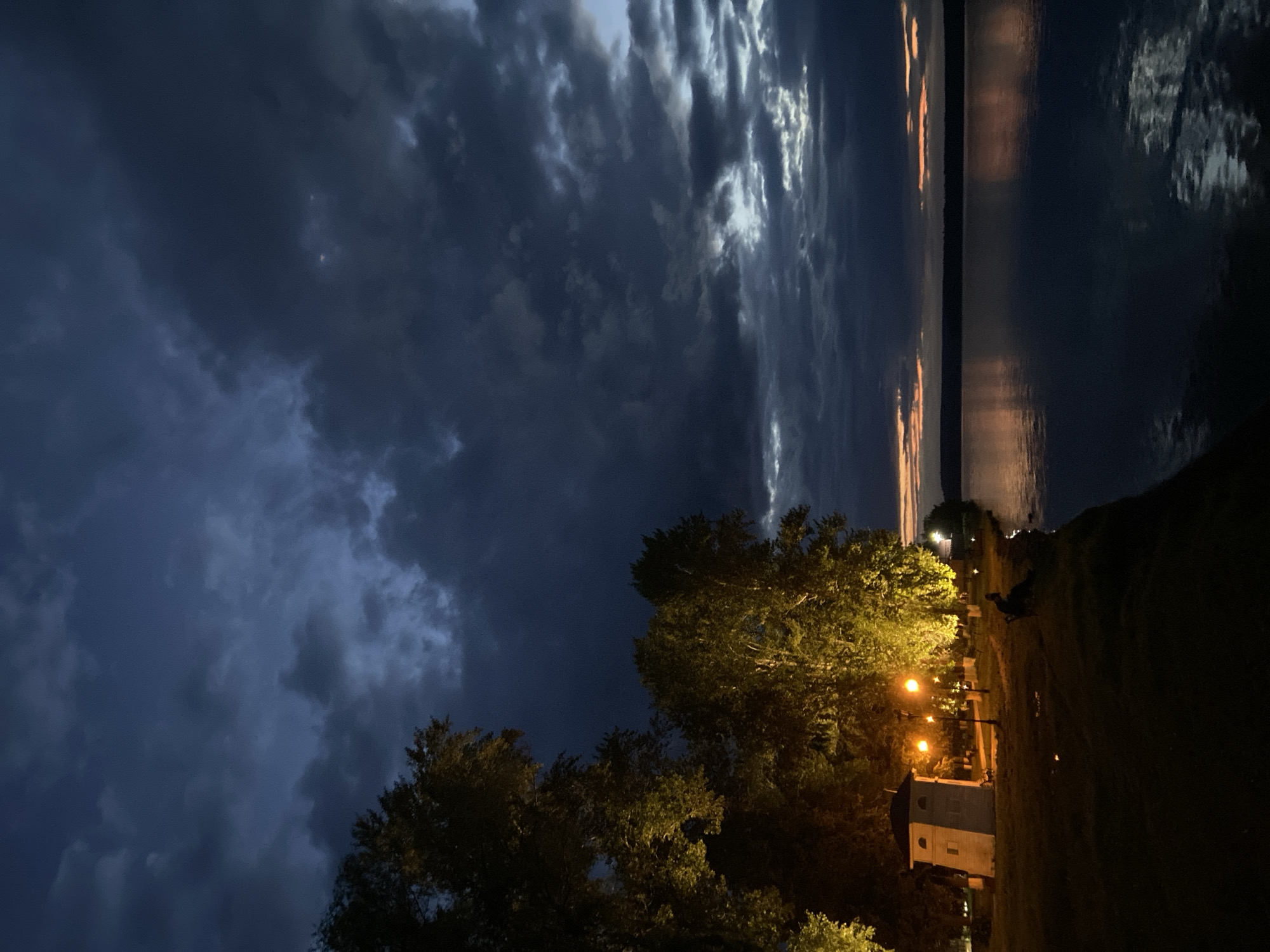 This image depicts a twilight scene by a body of water, likely the Votkinsk Reservoir, in Votkinsk, Russia. The sky is filled with dark, dramatic clouds, with hints of orange and pink from the setting sun visible near the horizon. The water's surface is dark and reflects the faint light from the sky.
On the left side of the frame, illuminated by warm streetlights, are several large trees with dense foliage. A small, white, lighthouse-like structure with a dark roof stands near the water's edge, also lit by streetlights. A few lampposts are visible, casting pools of light on the grassy area. A single person is seated on a bench near the water, silhouetted against the faint light. The ground is covered in grass and some patchy areas.
The overall atmosphere is serene and slightly melancholic, characteristic of dusk in a park or waterfront area. The image captures the transition from day to night in a natural setting within the city of Votkinsk.
