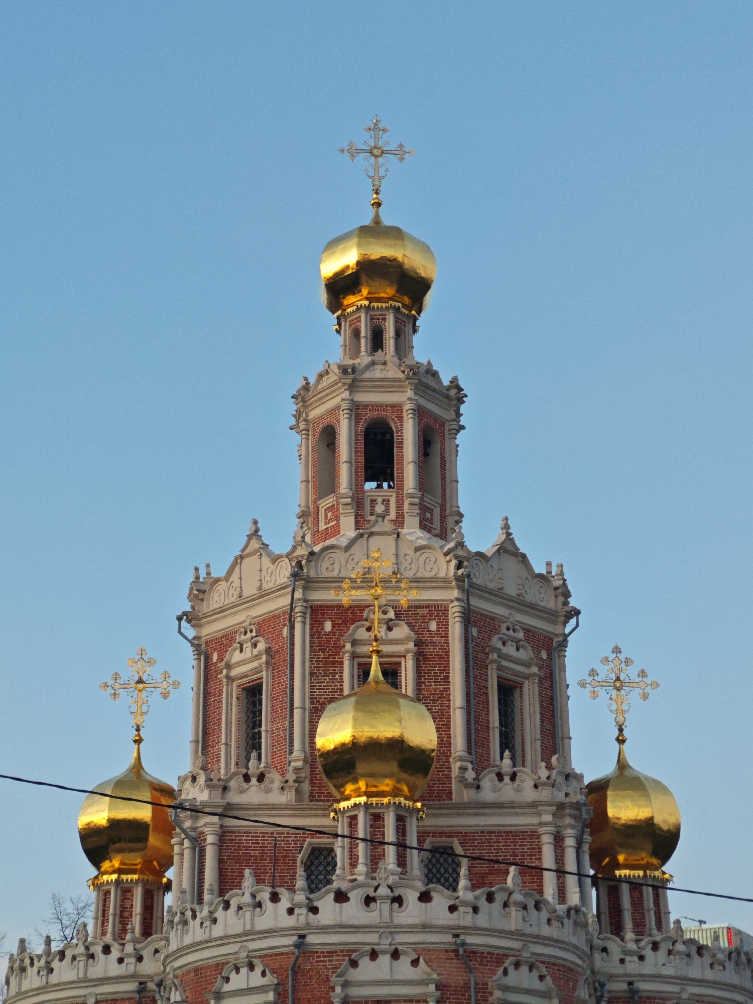 The image captures the Novodevichy Convent bell tower in Moscow, Russia. The tower is constructed of red brick and ornamented with white stone detailing. It is topped with five golden onion domes, each surmounted by a cross. The sky is a clear light blue. The image seems to be taken during the day.