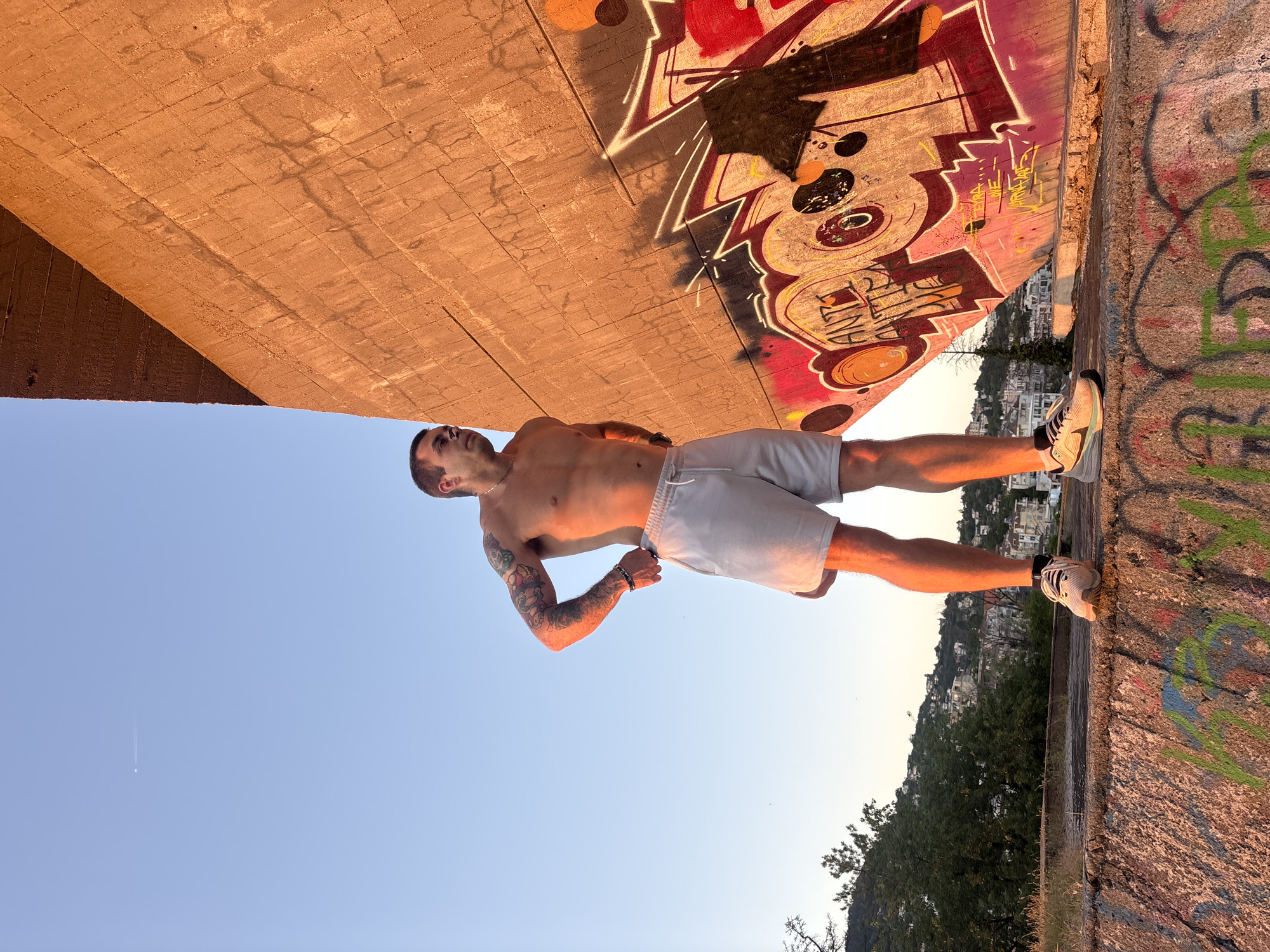 A shirtless man with tattoos on his arms stands on a concrete structure, looking out at a city. He is wearing light-colored shorts and athletic shoes. Behind him is a large concrete wall covered in graffiti. In the background, a cityscape with buildings and trees is visible against a clear blue sky. The scene is illuminated by the warm light of either sunrise or sunset, casting long shadows. The location is Varna, Bulgaria, specifically on the eastern side of the city near the Black Sea coast, possibly in an area with abandoned or repurposed military structures or industrial sites, given the concrete architecture and graffiti.