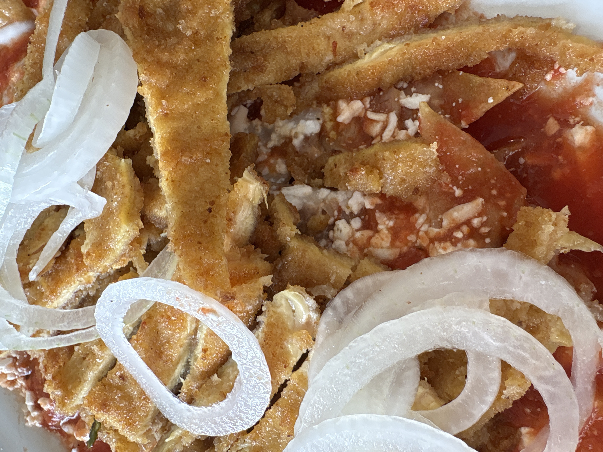 The image depicts a close-up of a Torta de Papa dish, a common street food in Mexico City, Mexico. The dish consists of fried potato cakes, sliced and layered, topped with red salsa, white cream or cheese, and raw onion rings. The texture of the potato cakes appears crispy on the outside and possibly soft inside. The salsa adds a red color contrast, while the white cream/cheese and onions provide visual and textural diversity.