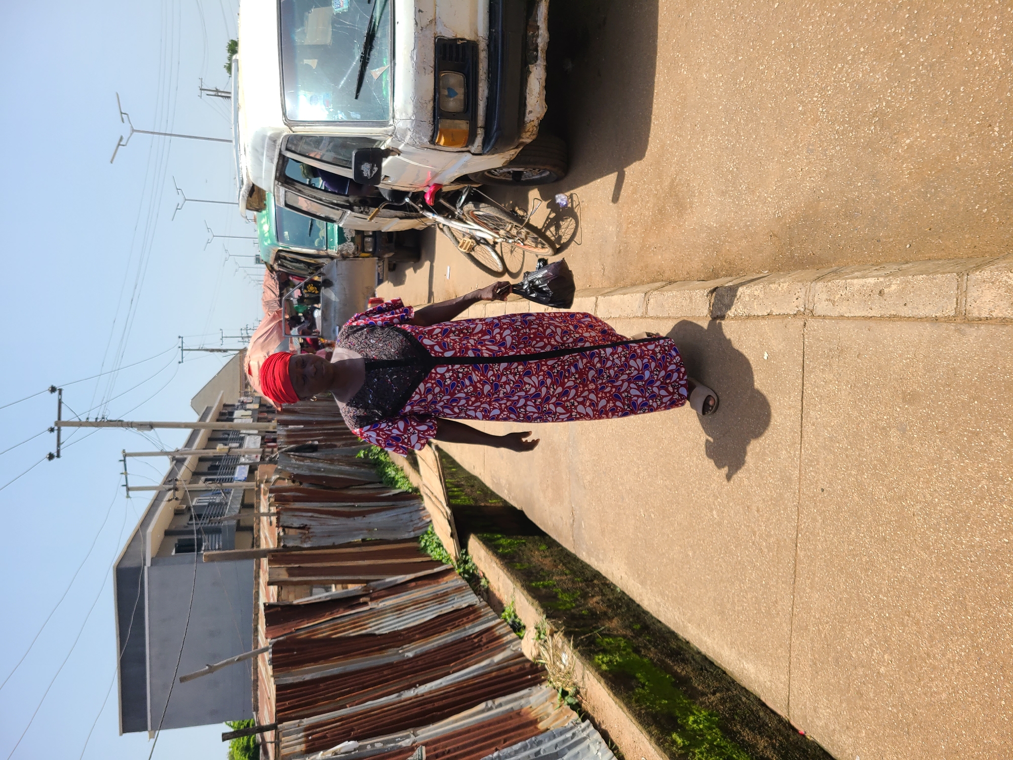 A medium shot captures a Black woman walking towards the camera on a paved sidewalk in Bukuru, Nigeria. She is wearing a red headscarf and a long, flowing dress with a red, white, and blue paisley pattern and a black lace trim around the neckline. She carries a black plastic bag in her right hand. To her left, a rusty corrugated metal fence lines a narrow, moss-lined drainage ditch. Beyond the fence, a gray, multi-story building with balconies and air conditioning units is visible, along with telephone poles and power lines. To the woman's right, several white and green minivans are parked along the street, with a bicycle leaning against the white minivan in the foreground. The street and sidewalk are dusty and a shadow of the woman is cast on the pavement. The sky is clear and blue.