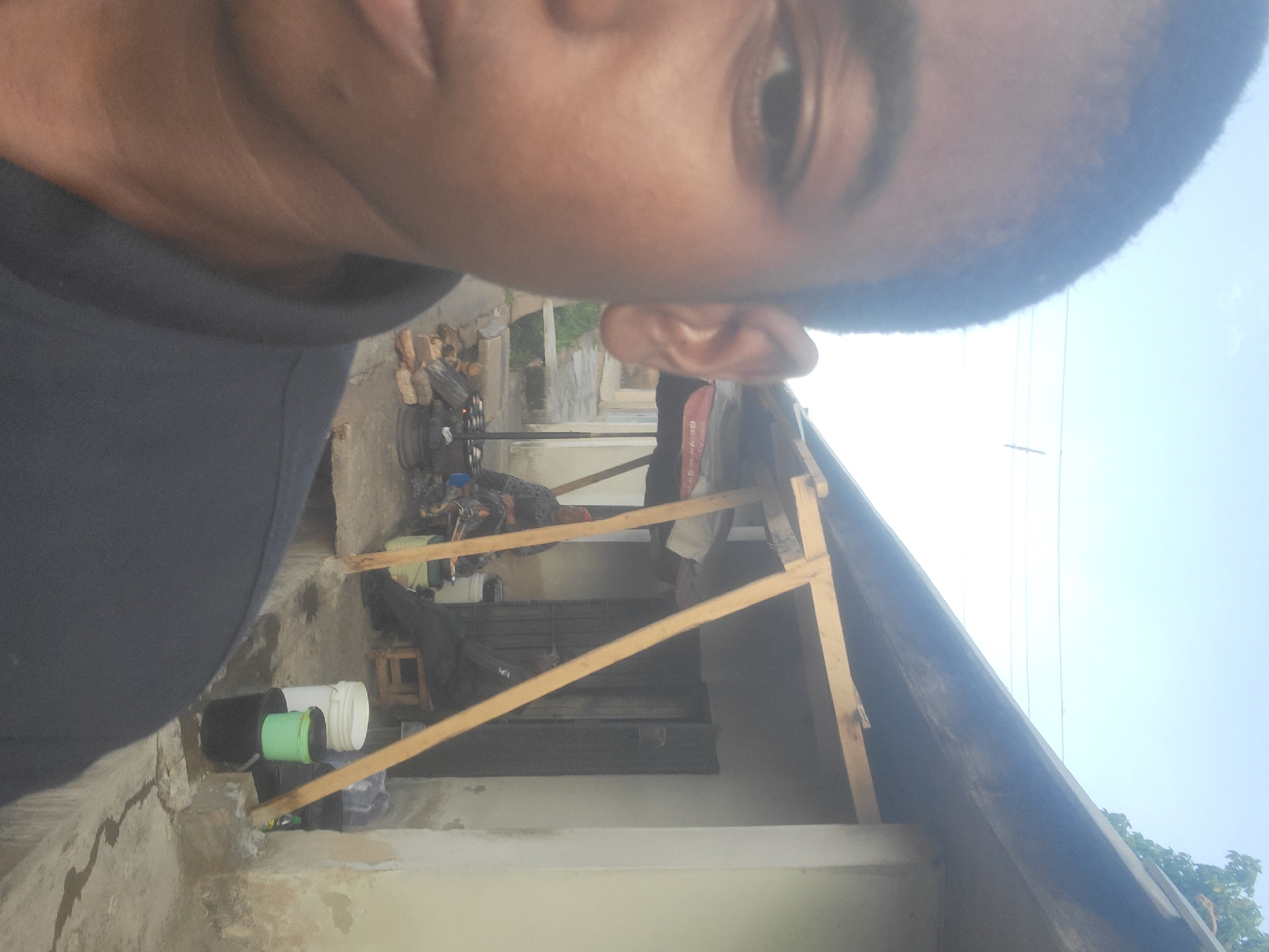 The image displays a partial selfie of a dark-skinned individual, likely male, occupying the left foreground, showing his upper torso, left eye, and part of his face. He has short, dark hair and is wearing a dark polo shirt. The background reveals an outdoor scene in what is identified as Jos, Nigeria. A light-colored, single-story building with a dark door is visible on the right. A makeshift wooden frame structure extends from the building, supporting a red and white canopy (partially visible, displaying "WWW.C..." and "080...") and providing shade. Two individuals are seated under this structure: one on the left in a patterned shirt, near an outdoor cooking setup comprising a metal pot on a stand over a fire with bricks; another individual, wearing dark attire, is seated on a low wooden stool further right, closer to the building's door. The ground is a paved surface, exhibiting wet patches. Several plastic buckets (white, green, black) and plastic bottles are scattered on the ground near the seated individuals. The upper portion of the image shows a clear blue sky with utility lines traversing it, and green foliage from a tree is visible above the building's roofline on the right.