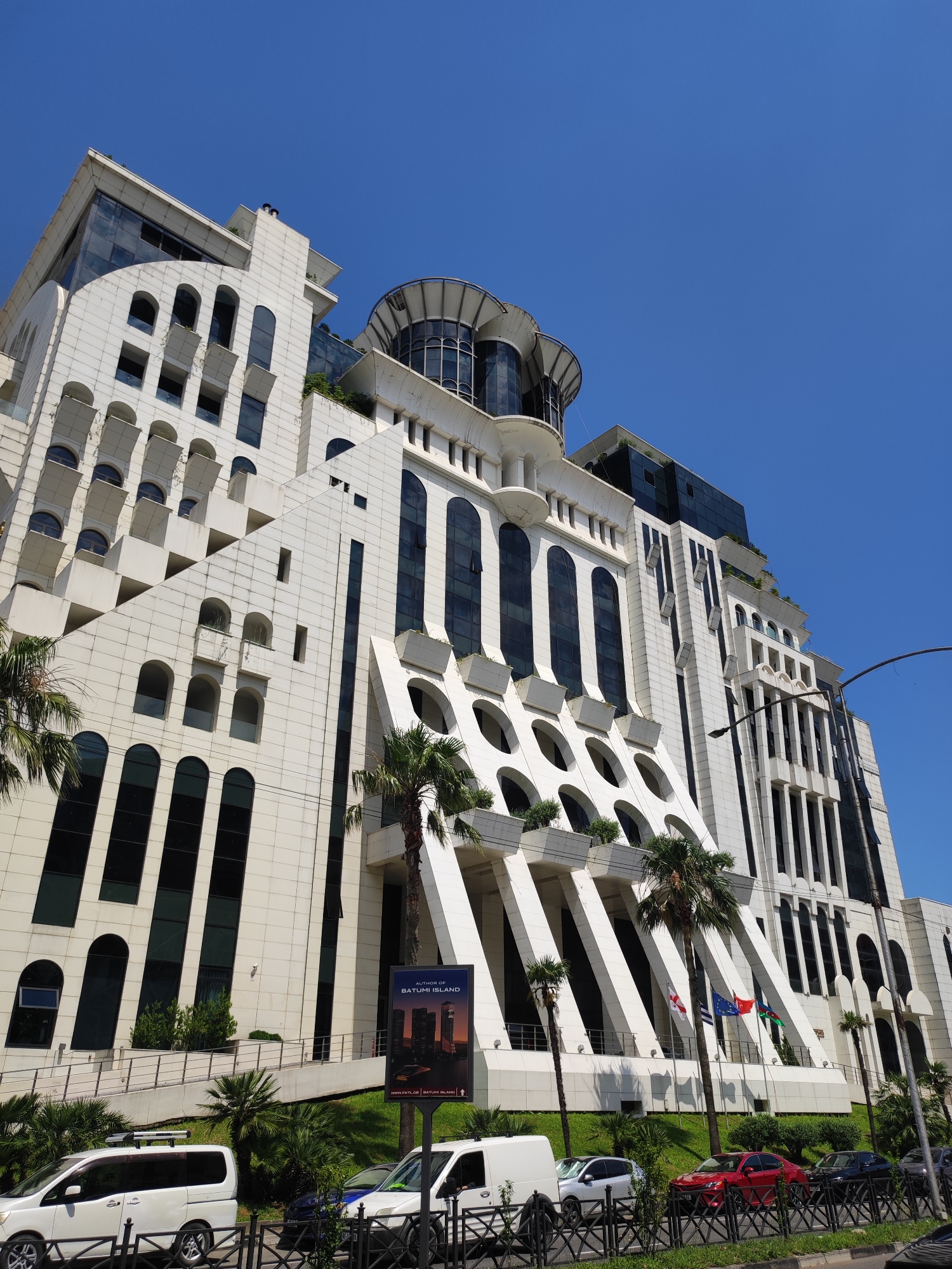 This is a low-angle, medium shot of the futuristic "Batumi Island" building in Batumi, Georgia, under a clear, bright blue sky. The building, constructed from white stone and glass, features a complex, stepped design with numerous arched windows and balconies. Slanted white supports create a dramatic visual effect on the lower levels. Palm trees are visible in the foreground, and a street with cars and a black metal fence runs along the bottom of the image. A billboard for "Batumi Island" is positioned to the right of center, displaying a nighttime cityscape. Flags of various nations are planted in front of the building.