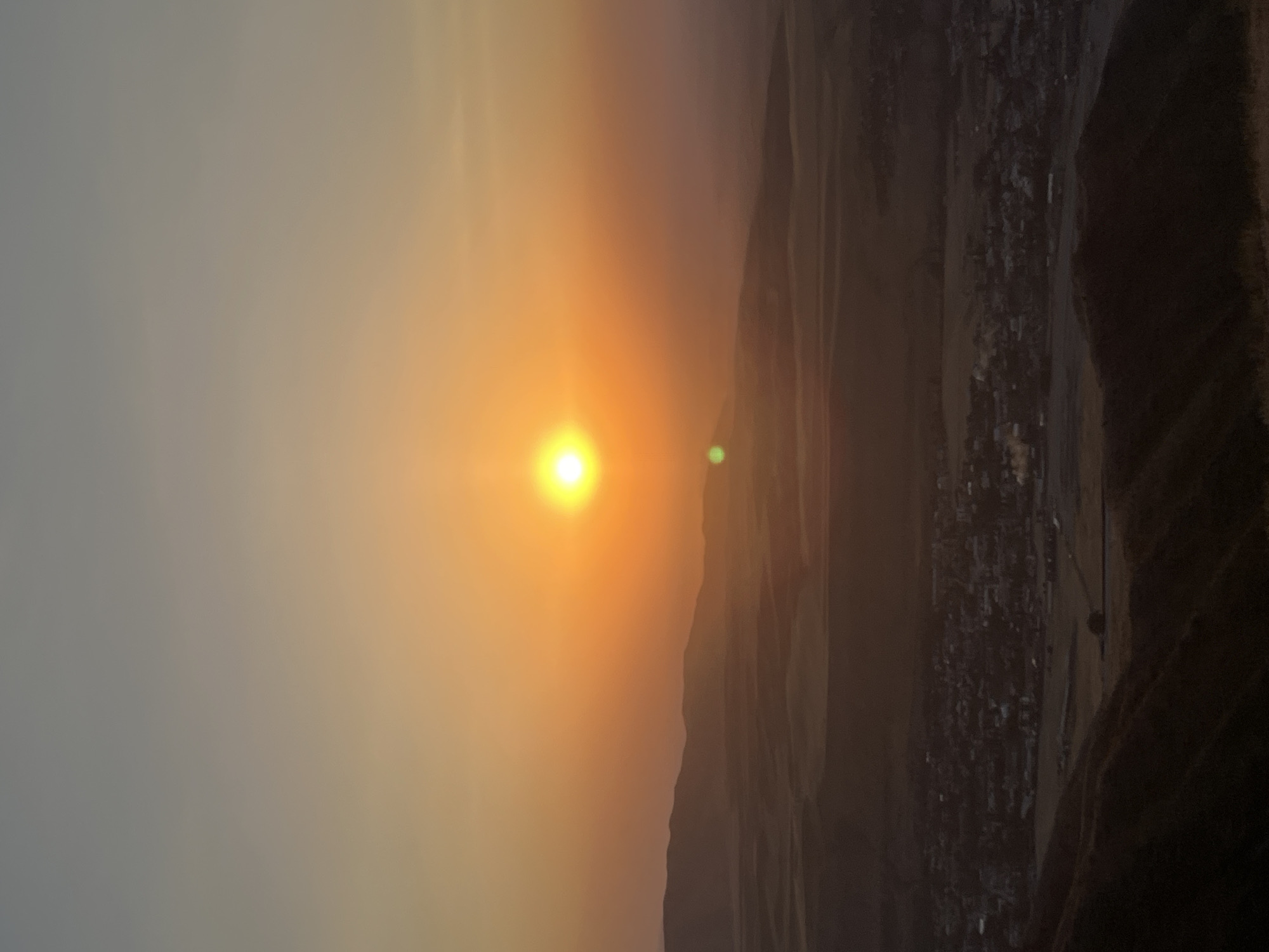 A high-angle, vertical-format, photograph captures a sunset over a city and its surrounding landscape. The sun, a bright orange orb, is centrally located in the upper half of the image, casting a warm glow that transitions from yellow near the sun to orange and then to a hazy gray at the top of the frame. Below the sun, distant hills create a silhouetted horizon, their forms softened by the atmospheric haze.

In the middle ground, a small city is visible, characterized by clusters of buildings and a network of streets. The city appears to be nestled within a valley or plain, with fields or agricultural land surrounding it. The foreground is dominated by the dark, textured slopes of hills, suggesting the photograph was taken from an elevated viewpoint, possibly a mountain or a high ridge overlooking the city. The overall atmosphere is serene and slightly hazy, typical of sunset conditions. The location is identified as Besh Kungei, Kyrgyzstan.