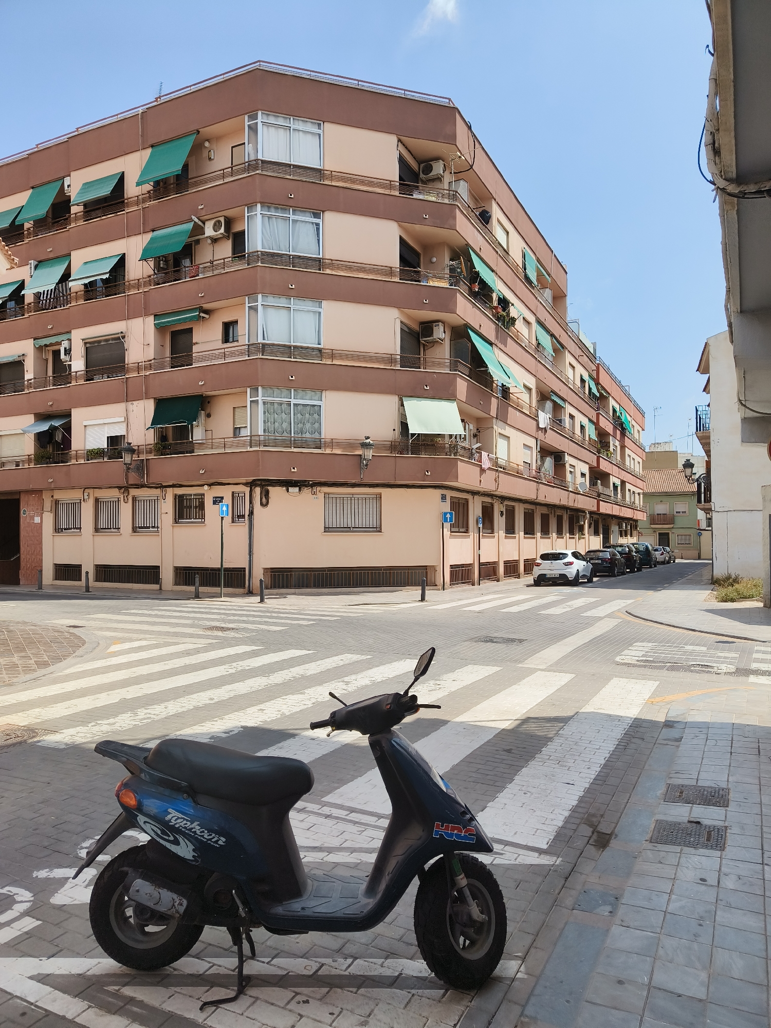 This is a street-level, eye-level, medium shot captured on a bright, sunny day in the Benimàmet-beniferri area of Valencia, Spain. The scene is dominated by a large, multi-story apartment building with a beige and brown facade. The building features multiple balconies with green awnings extended over them, and several windows are visible, some with curtains drawn. Air conditioning units are mounted on the exterior walls. In the foreground, a dark blue scooter is parked on a pedestrian crossing. The scooter has "Typhoon" and "HRC" branding visible. The crossing is marked with white stripes on a paved surface, and beyond it, a street with several parked cars stretches into the distance. The sky is a clear blue with a few wispy clouds. The overall impression is of a typical urban residential street.
