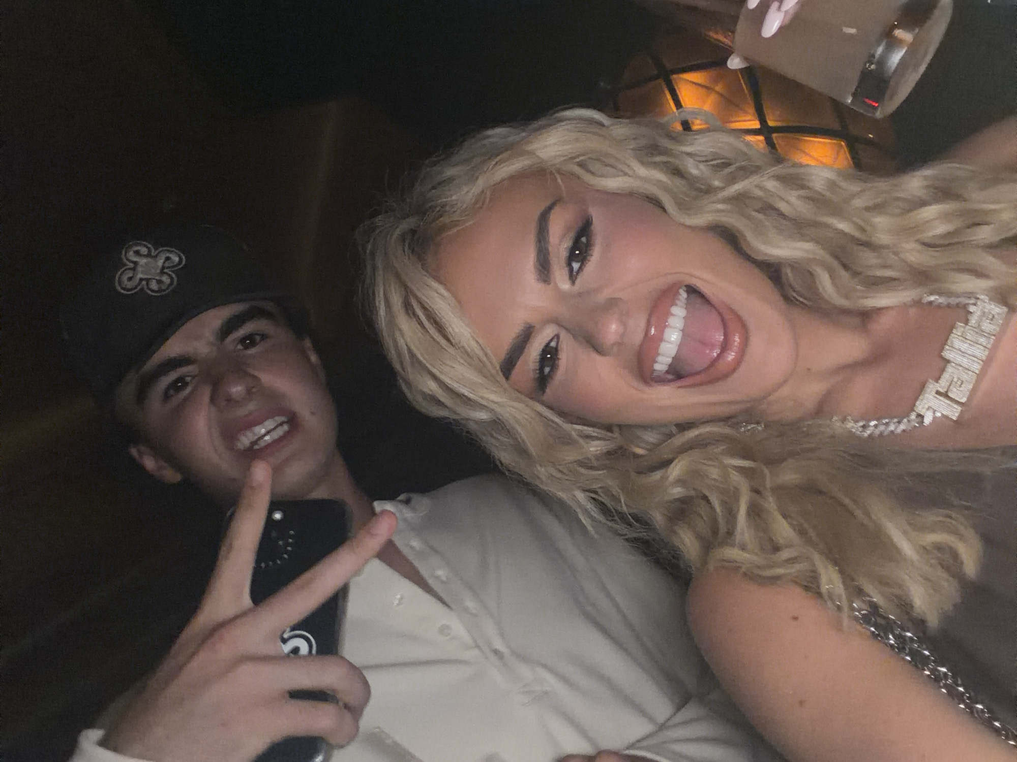 Two people pose for a selfie in a dimly lit interior that may be a bar or nightclub in Miami Beach, United States of America. On the left is a young man with dark hair wearing a dark baseball cap. He is holding up his left hand in a peace sign and holds a dark phone in that hand. He wears a cream-colored button-down shirt. On the right is a woman with long blonde, wavy hair. She wears a chain necklace with the name "Talia" on it. She is smiling widely and has her mouth open. There is a glass in the upper-right corner. The background is dark with some illuminated square patterns.