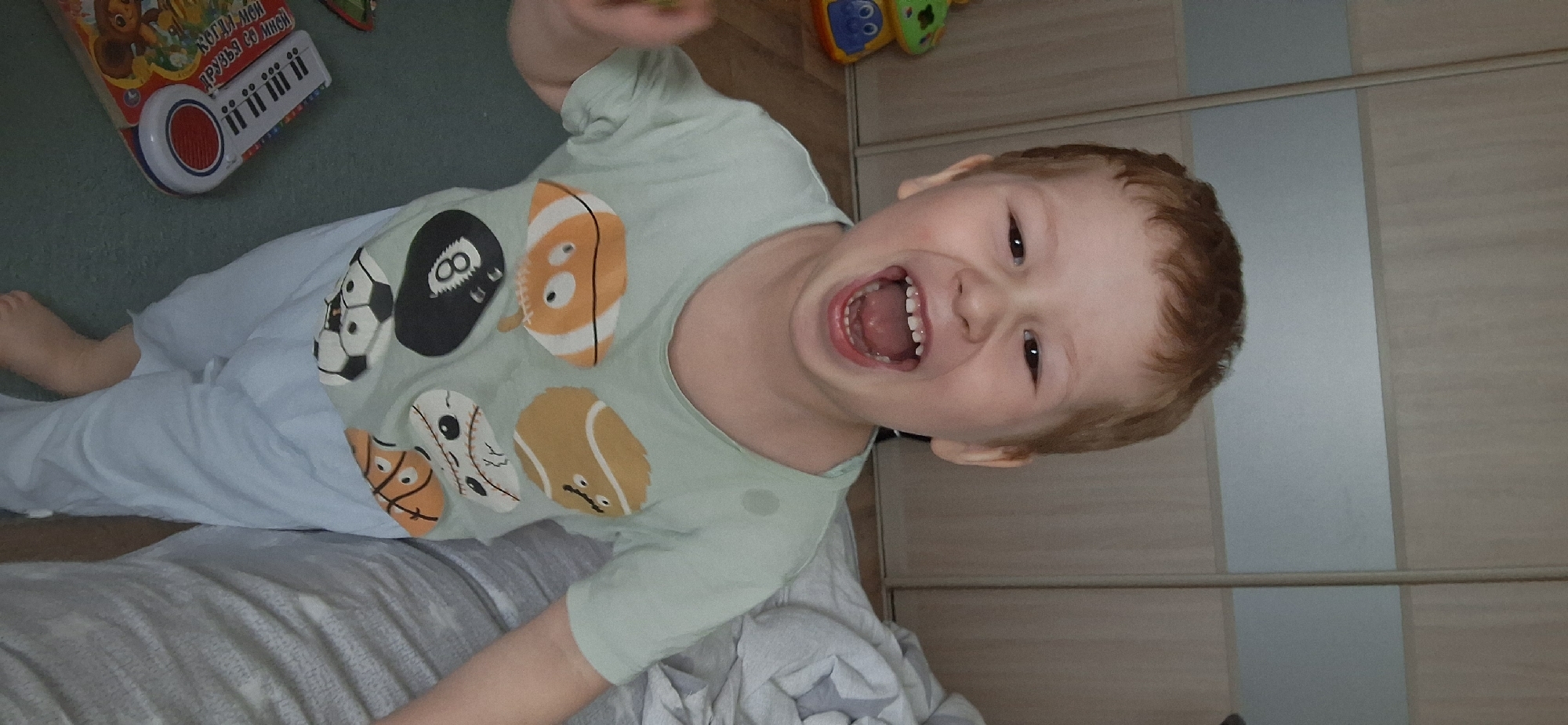 The image depicts a young, light-skinned child with reddish-brown hair, lying on what appears to be a bed covered with a gray and white patterned blanket and white sheets. The child is wearing a light green t-shirt with various cartoonish sports ball designs, and light blue pants. The child is looking at the camera, mouth open, expressing a joyful or excited emotion. In the background, to the left, there is a blue and green floor, with a colorful toy on it. To the right, there is a light-colored, wooden-paneled wall. Given that the location is Chaikovsky, Russia, this is likely a domestic setting, possibly the child's bedroom.