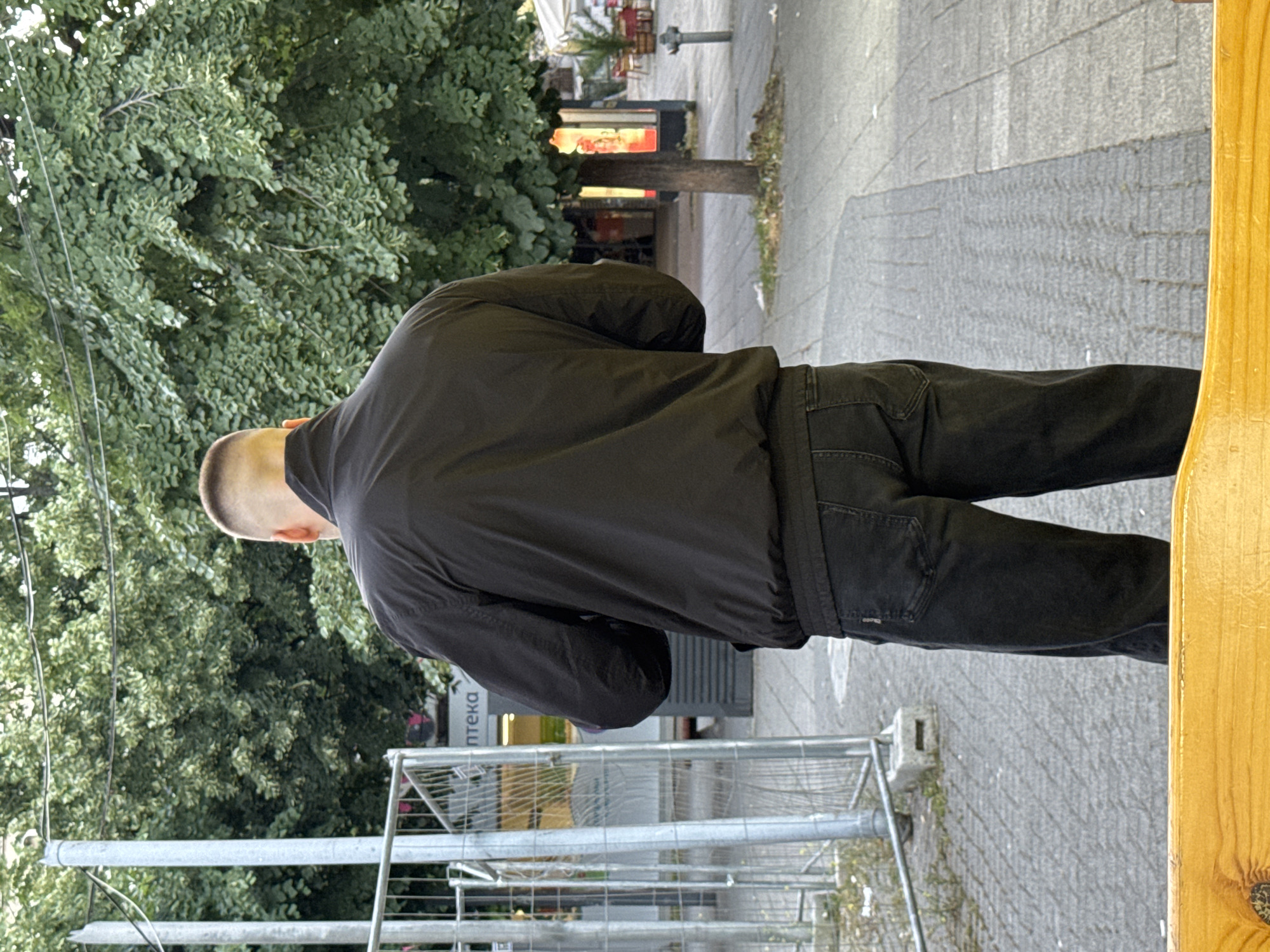 A man with a very short haircut stands facing away from the camera, wearing a black jacket and black jeans. He is positioned on a paved walkway, with a metal fence to his left and large trees in the background. A wooden bench is visible in the foreground. The scene appears to be in an urban setting. The image was taken on Aleko Bogoridi Street in Burgas, Bulgaria.
