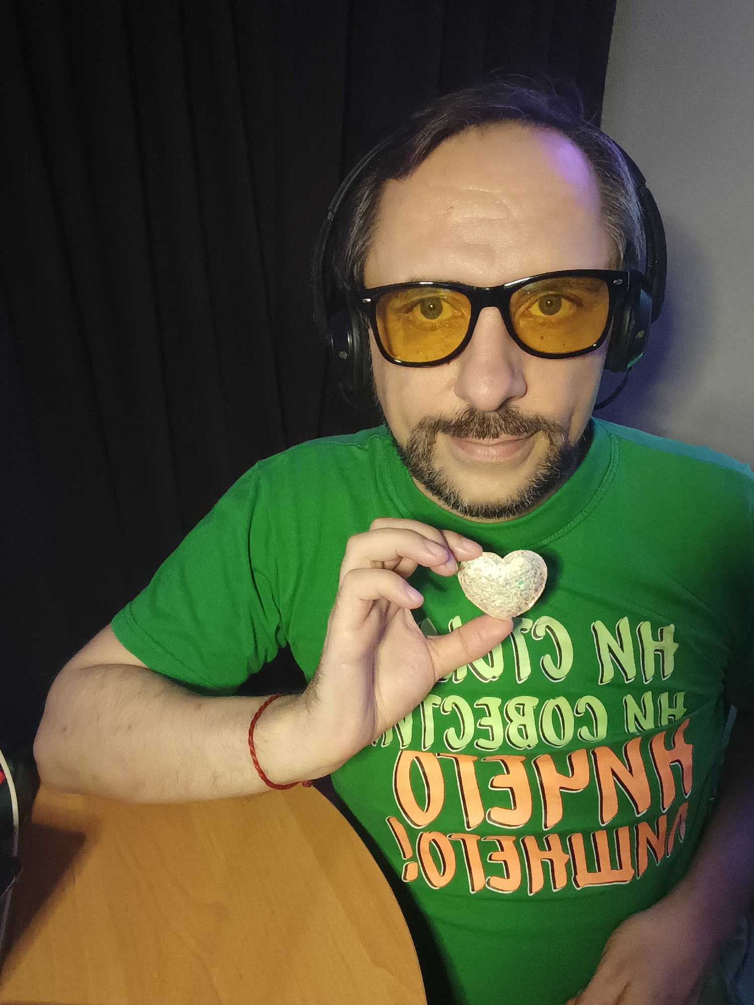 A man with a goatee, wearing headphones and yellow-tinted glasses, is shown from the chest up, holding a heart-shaped object. He is wearing a green t-shirt with an orange and green slogan. The man's left arm rests on a wooden table. A red string bracelet is visible on his left wrist. The background consists of a dark, pleated curtain. The image was taken indoors.