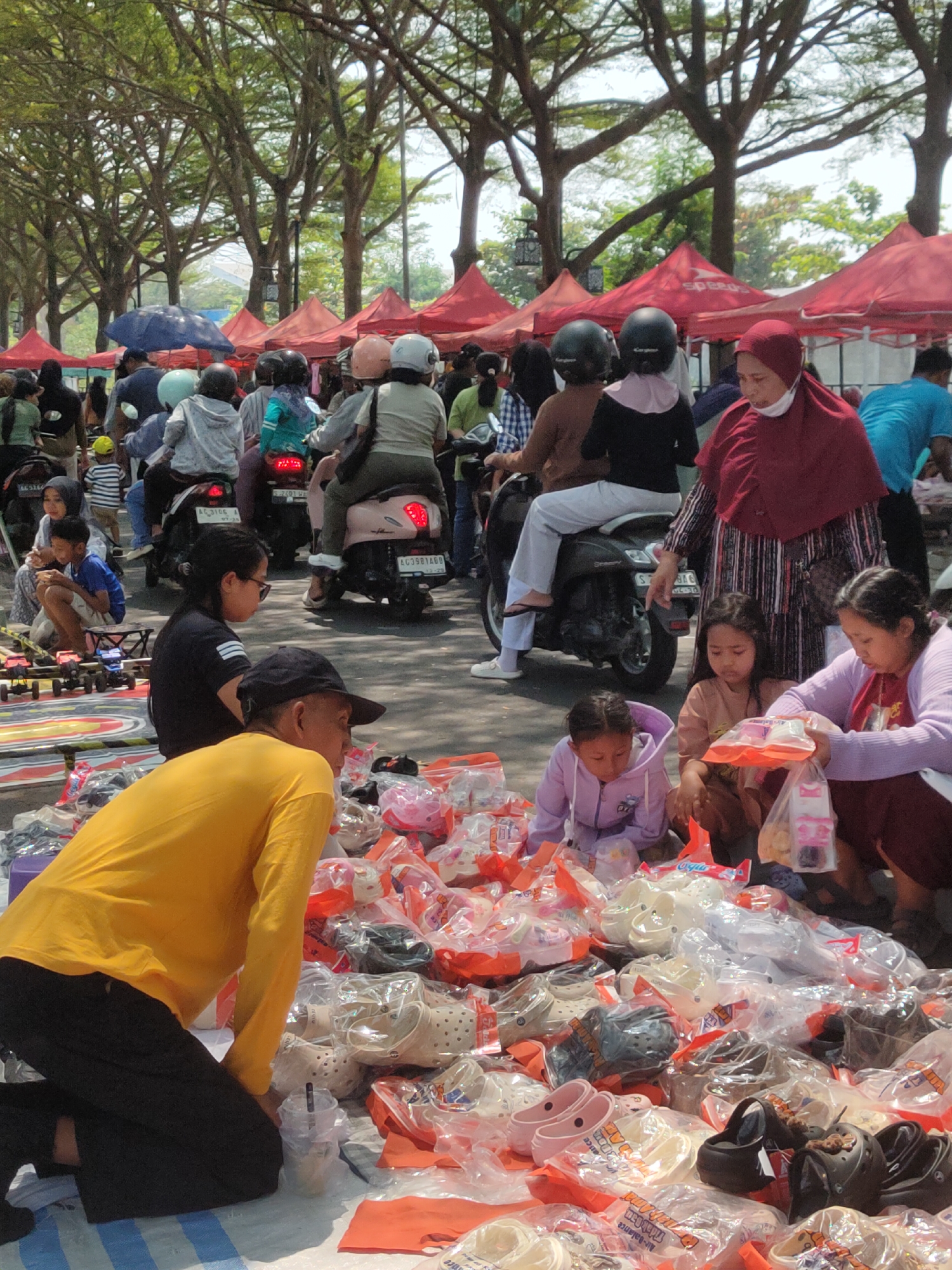 The image depicts a busy outdoor market scene in Ngasem, Indonesia, likely on a sunny day. The foreground is dominated by a large pile of shoes, primarily clogs, enclosed in clear plastic packaging, spread across an orange tarp on the ground. A man in a yellow long-sleeved shirt and black pants is kneeling in front of the shoes, facing away from the camera. Behind him, a woman in a purple cardigan and a young girl in a purple hoodie are kneeling and examining some of the packaged items. Another young girl in a light brown shirt sits next to them.
Further back, a bustling crowd is visible, with many people on motorcycles. Red tents and umbrellas line the street, indicating vendor stalls. People are wearing casual clothing, and several individuals are wearing helmets. A woman in a maroon headscarf and a face mask stands near a motorcycle. The background shows trees with dense foliage, suggesting a location with ample greenery. The overall atmosphere is one of a lively market with commerce and pedestrian traffic.