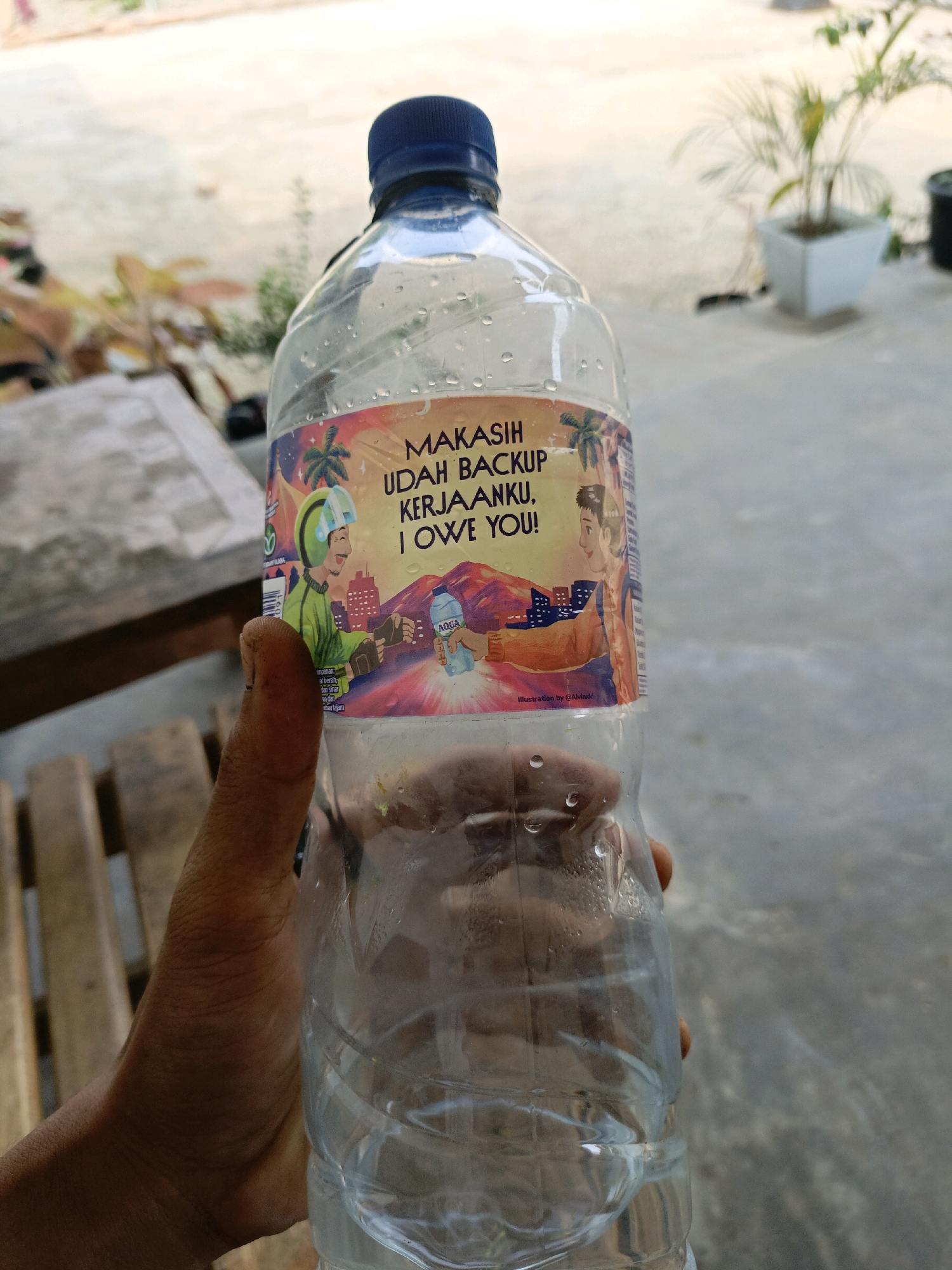 A close-up, eye-level, outdoor shot shows a person's hand holding a clear plastic bottle of AQUA mineral water. The bottle has a blue cap and a colorful label with text that reads "MAKASIH UDAH BACKUP KERJAANKU, I OWE YOU!". The label also features an illustration of two people, one in a green uniform with a helmet, and another person reaching out to hand a bottle of AQUA water. The background consists of a light-colored, mottled ground, possibly concrete, with hints of vegetation and a potted plant in the distance. The lighting suggests it is daytime, with shadows indicating sunlight. The specific location in Kota Agung, Indonesia, is not identifiable from the visual cues in the image.