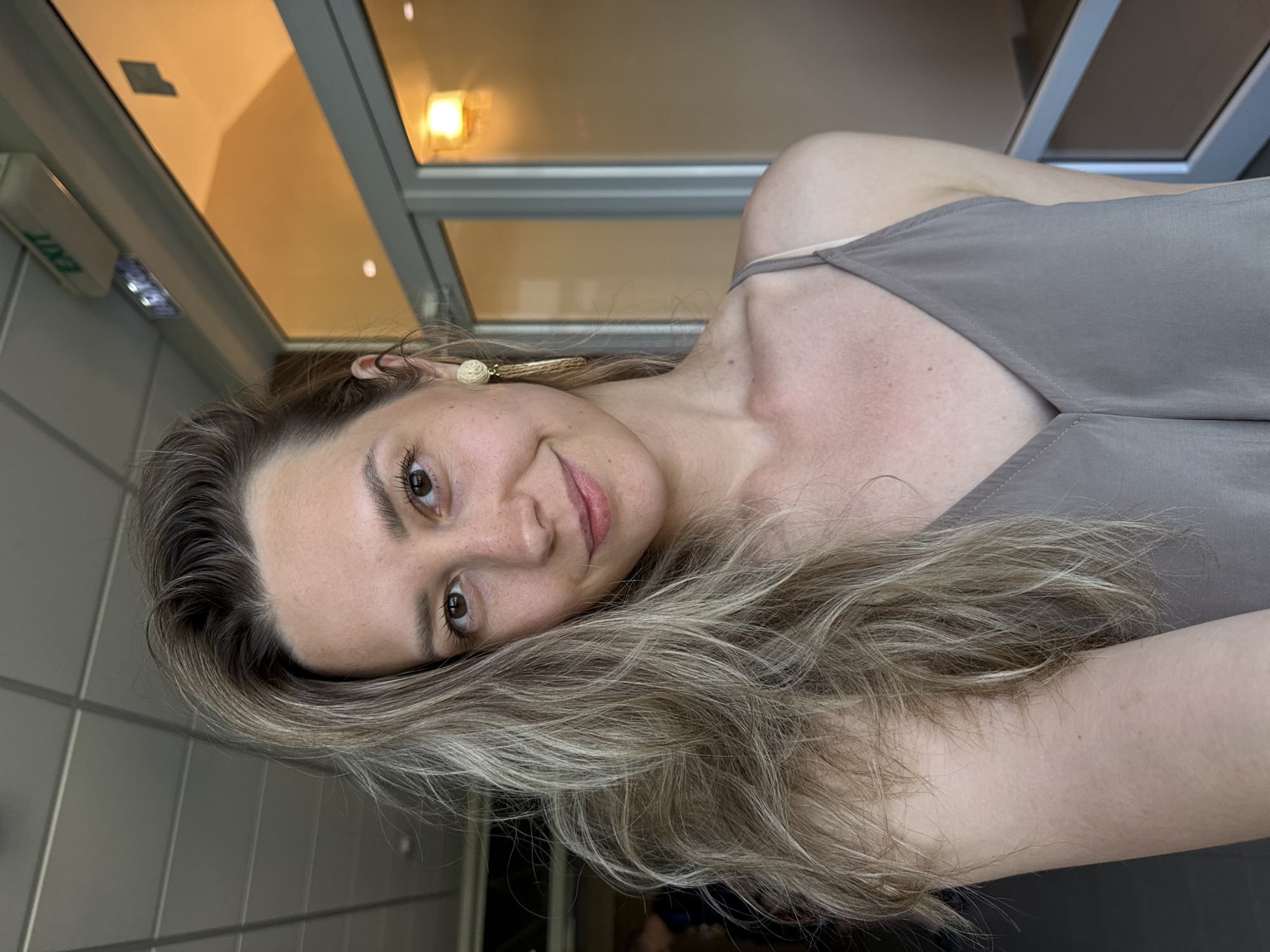 A close-up selfie shows a young woman with blonde, wavy hair and brown eyes. She is wearing a beige spaghetti strap top and a gold, tassel-like earring in her right ear. Her left cheek is slightly flushed, and she has freckles across her nose and cheeks. She is looking towards the camera with a slight smile. Behind her, a hallway with a tiled ceiling is visible, along with a glass door that leads to an area illuminated by warm lighting. An "EXIT" sign is mounted on the ceiling above the doorway. The overall setting appears to be an indoor public space, possibly a hotel or administrative building, given the "EXIT" sign and polished interior. The specific location is the city of Nebug, Russia, as indicated by the metadata of the original image file.