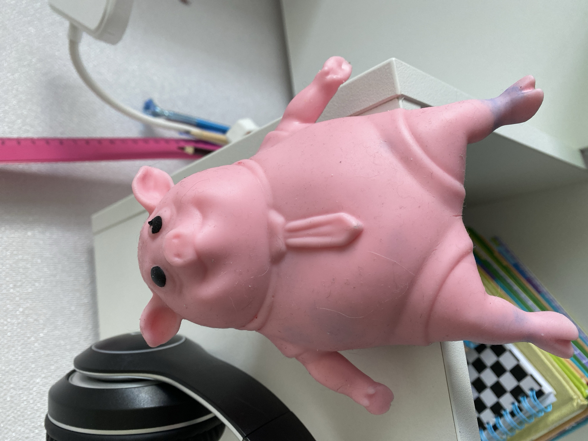 The image shows a close-up of a pink rubber pig toy positioned on the edge of a white shelf. The pig is lying on its side, with its head tilted slightly upwards, revealing two black eyes and a snout. Its limbs are splayed out, with one hind leg showing a purple discoloration towards the foot.
In the background, a white object, possibly a desk lamp or power adapter, is visible, connected to a white cord. Below the pig and to the left, the top of a pair of black headphones is visible. Behind the pig and to the right, a stack of colorful items, including notebooks with a checkered pattern and what appear to be pencils or pens, are neatly arranged on the shelf. A pink ruler is also visible in the lower left quadrant of the image, lying horizontally. The overall scene appears to be an indoor setting, likely a desk or workspace, with the lighting suggesting it is daytime. There is no discernible text in the image.