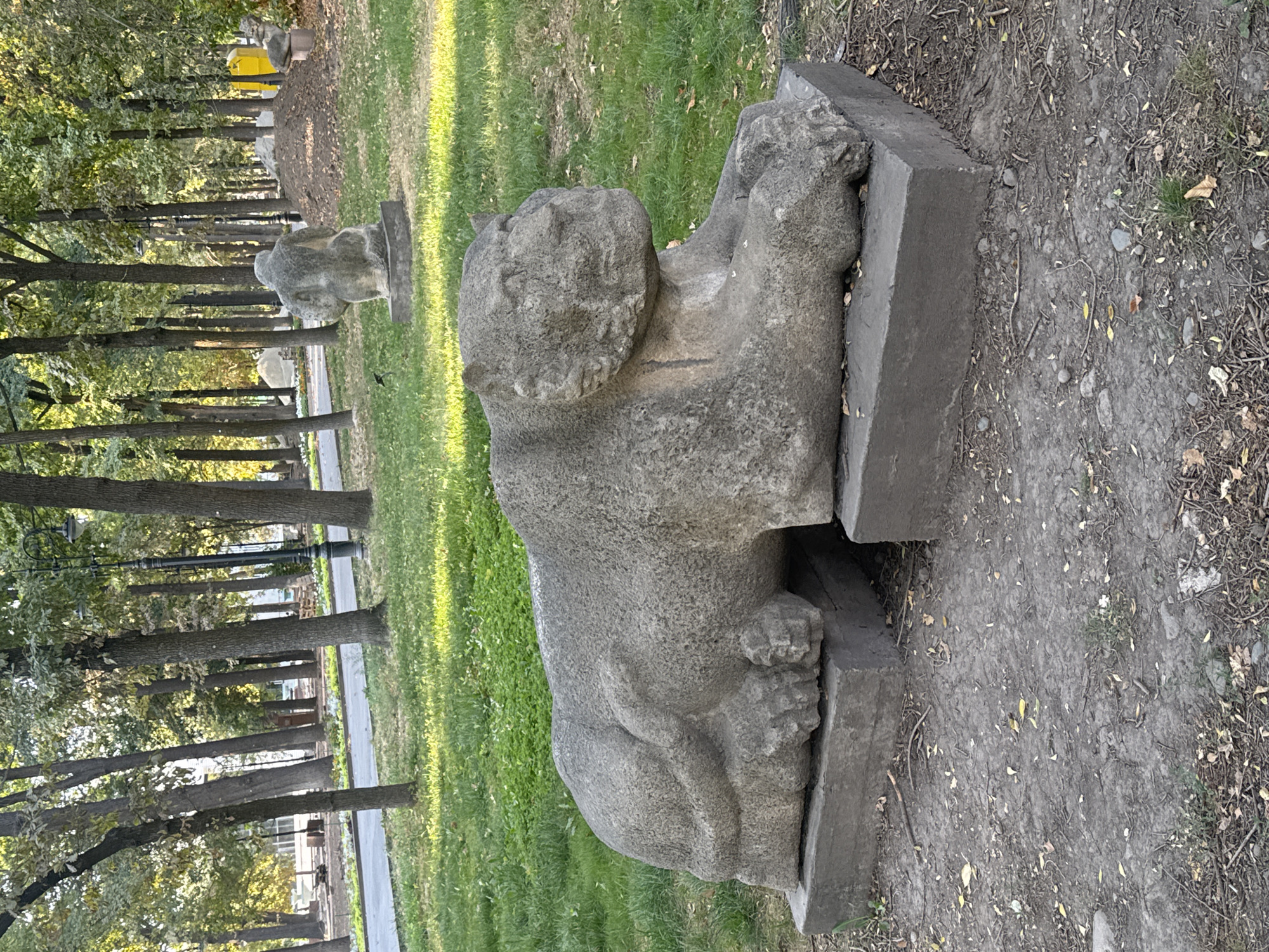 The image shows a stone sculpture of a lion in a park-like setting. The lion sculpture is in a reclining position and is placed on two rectangular stone blocks. The sculpture is weathered and appears to be ancient. The background consists of tall trees with green foliage, a paved path, and a lamppost. In the distance, another stone sculpture is visible, which appears to be of a human figure. The ground is covered with grass and fallen leaves. This scene is located in Oak Park, a prominent green space in Bishkek, Kyrgyzstan, known for its collection of Soviet-era sculptures and monuments.