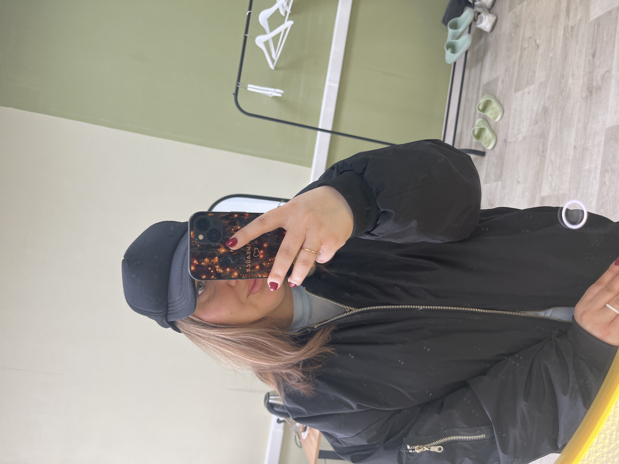 A mirror selfie, taken from a slightly elevated angle, features a person partially visible in the frame, wearing a black puffer or bomber jacket and a black baseball cap. They are holding a smartphone with a distinctive case up to capture their reflection. The person has light brown or blonde hair, a small portion of their face including one eye and part of their lips is visible, and they are wearing dark red nail polish and a gold ring on their right hand's ring finger.
The phone case is black with a pattern resembling warm, twinkling lights or stars, and visible mirrored text that reads "РОДАЯИЯ" (which would be "ЯИЯАДОР" unmirrored) along with a heart symbol. Underneath the jacket, a light blue or grey top is visible.
The background, seen in the mirror's reflection, consists of a room with two-toned walls: the upper portion is a light sage green, and the lower portion is off-white or cream. The floor is covered with light-colored wooden laminate. To the left, a black metal clothing rack with several white hangers is visible. On the floor, near the wall, there are two pairs of slippers: one pair of light mint green slides and another pair of light yellow-green slides. A white sneaker or shoe is also visible on the floor near the wall. The overall setting appears to be an indoor space, possibly a room or a changing area.