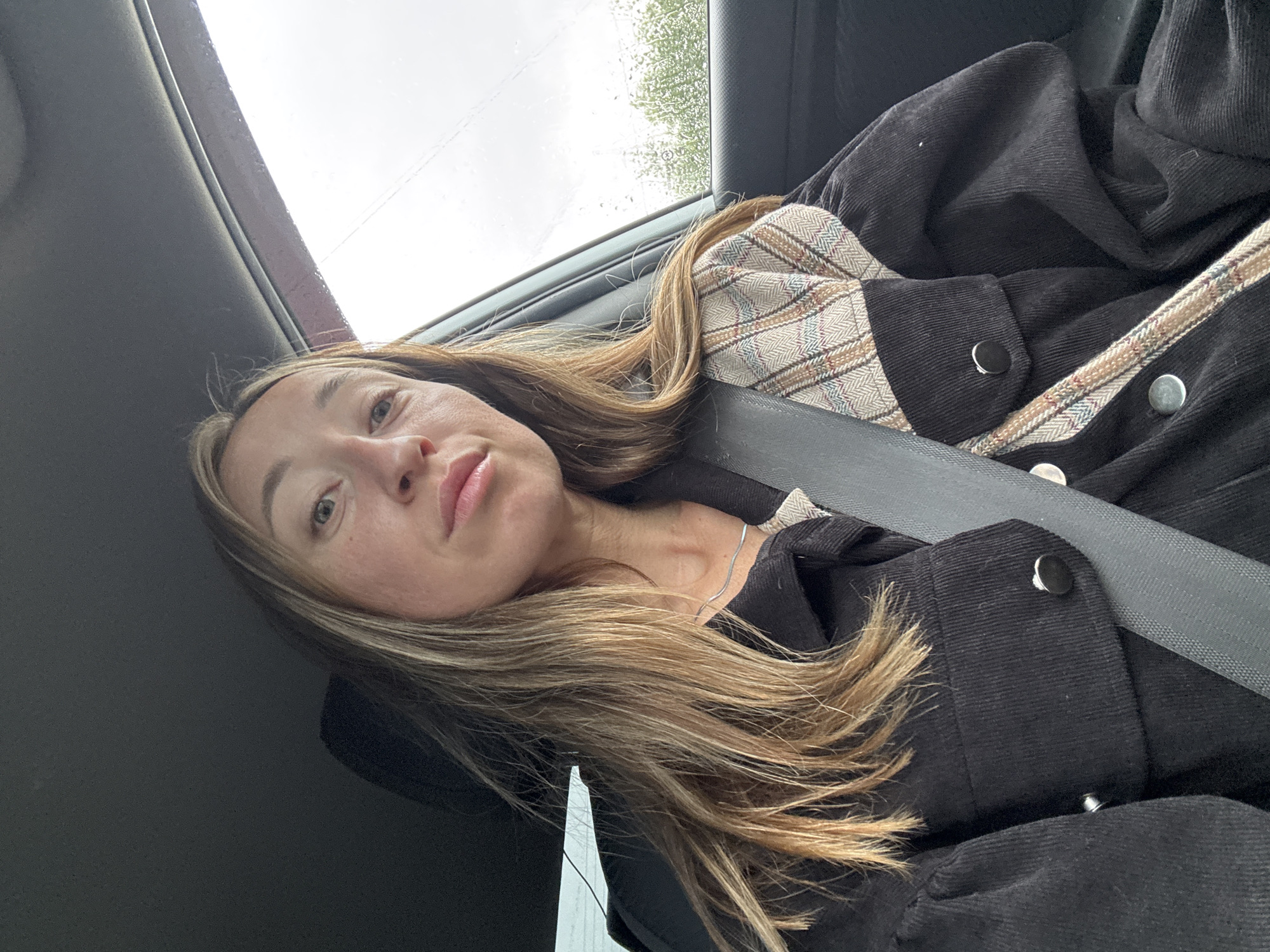 A woman with long, blonde hair is sitting in the driver's seat of a car. She is wearing a black jacket with a plaid detail on the shoulders. A seatbelt is visible across her chest. The window of the car is open and the weather appears to be overcast with rain. The background shows trees and foliage, suggesting an outdoor, possibly rural or suburban, setting. The woman is looking directly at the camera.