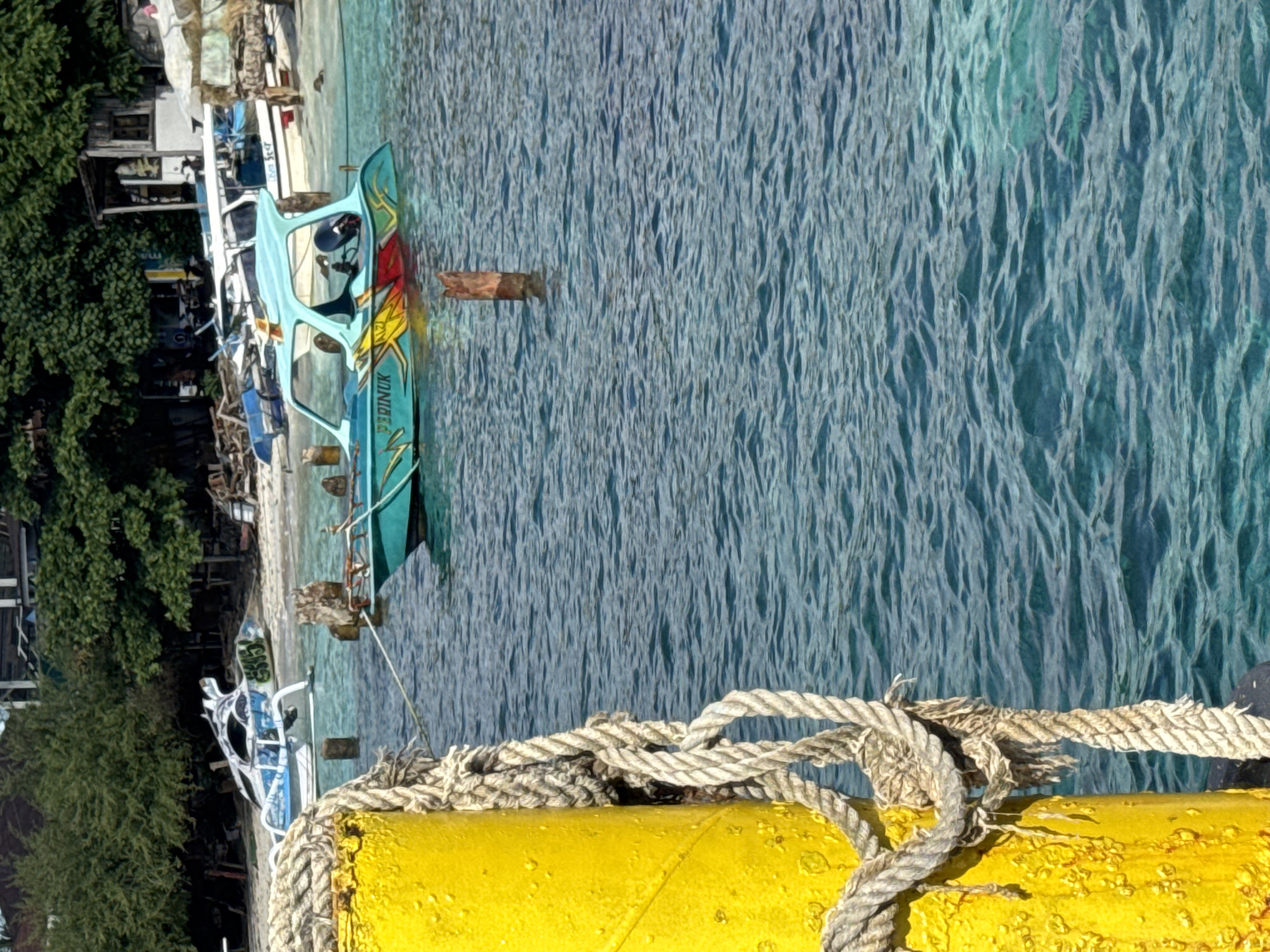 A vibrant coastal scene, likely in Indonesia, is captured from a slightly elevated perspective, possibly a pier. In the foreground, a bright yellow cylindrical object, possibly a buoy or part of a dock, dominates the lower right, with thick, weathered rope loosely wrapped around it.
Beyond this foreground element, clear, shimmering turquoise water extends across the frame, exhibiting distinct ripples from a gentle breeze or current. Several boats are present, either moored or beached along a sandy or rocky shoreline that borders lush green vegetation and rustic wooden structures on the left.
The most prominent vessel, positioned centrally, is a small, light blue boat adorned with eye-catching red and yellow artistic designs on its side. The word "PERINUK" is clearly visible in white capital letters on its hull. This boat appears to be a traditional longtail or outrigger-style boat, common in the region. Other smaller boats, including a white one, are partially visible further back along the shore.
The scene is bathed in bright daylight, suggesting a sunny day, with no clouds visible. The water's clarity indicates shallow depths near the shore. The overall impression is one of a peaceful yet active tropical waterfront. No individuals are explicitly visible.