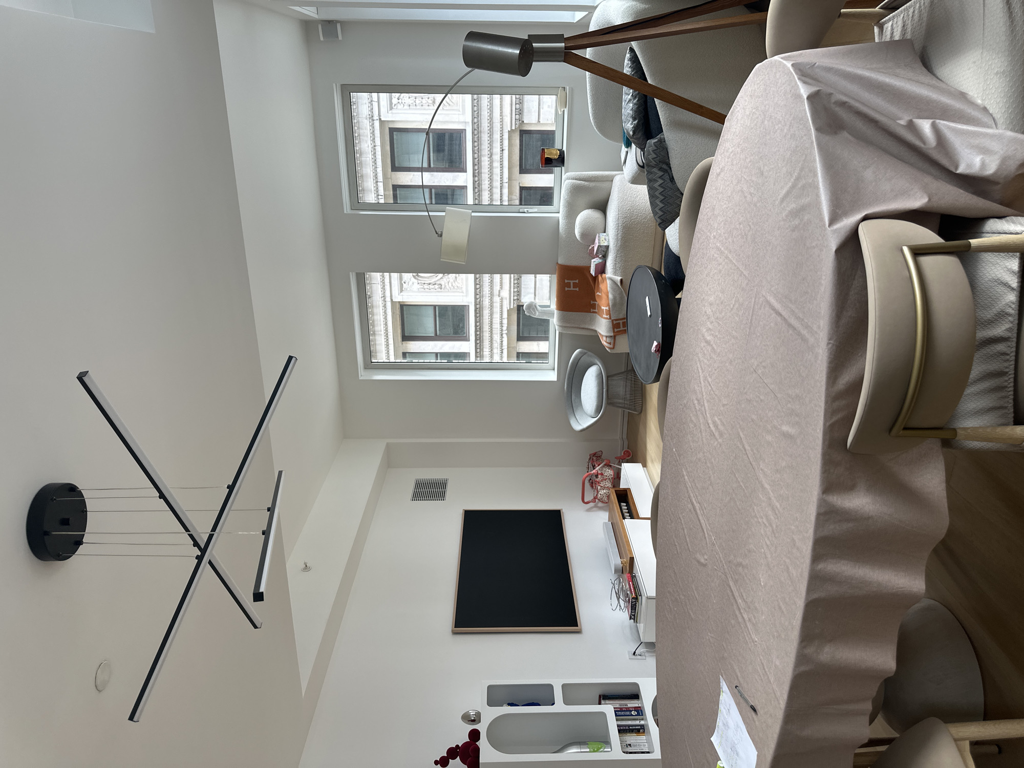 The image shows a well-lit interior space, likely an apartment or condominium, in New York City, United States of America. The space is furnished with modern pieces, including a sofa, chairs, and a dining table covered with a large beige cloth. Two large windows offer a view of a classic building exterior with multiple windows. The inside features a modern chandelier, a large black framed object, likely a TV or chalkboard, on the wall, a white shelving unit, and a floor lamp near the window. The room is tidy and well-maintained, suggesting a comfortable and contemporary living space.