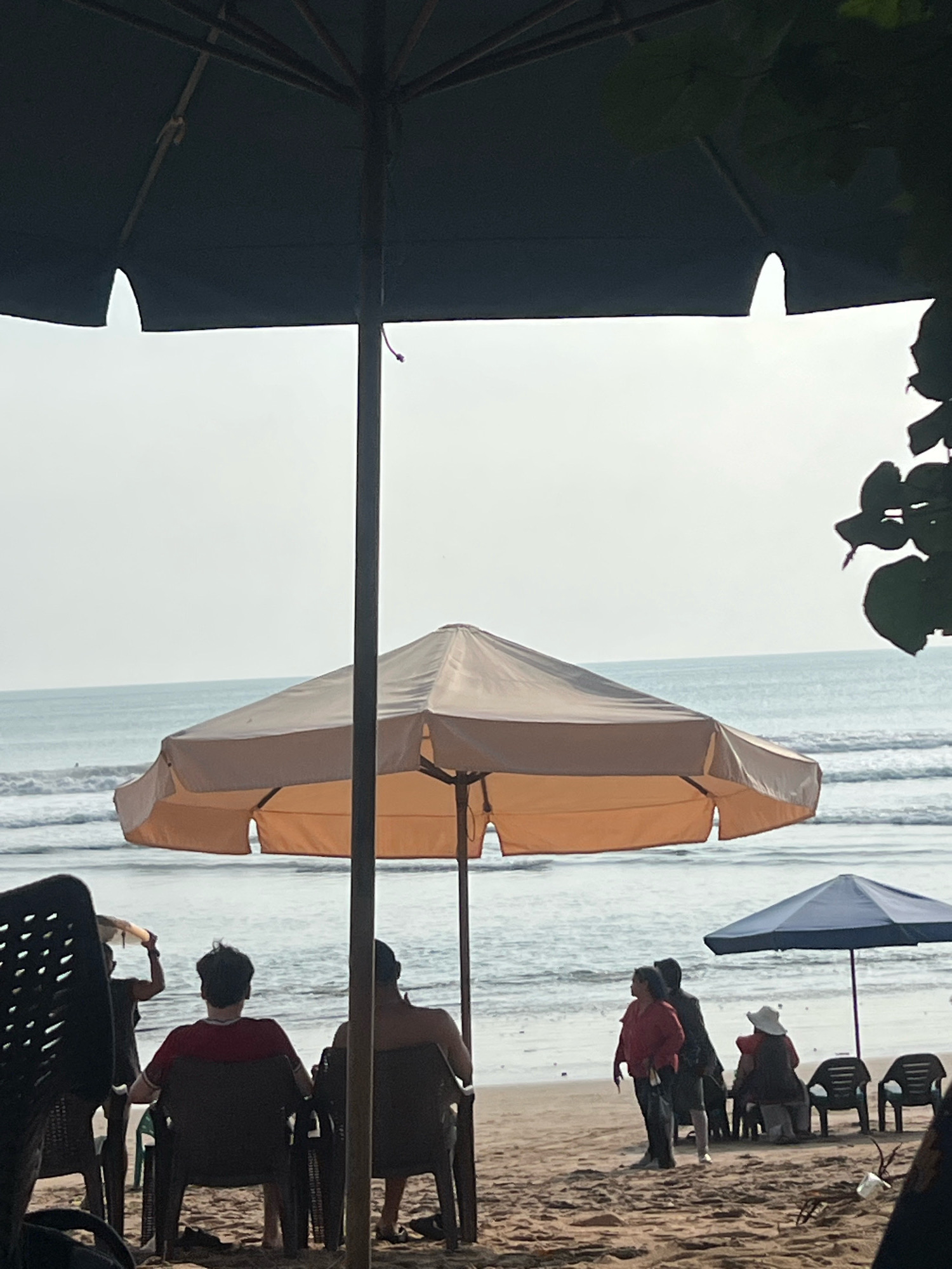 A beach scene in Kuta, Indonesia, captures a cloudy day with a calm ocean. In the foreground, two large umbrellas provide shade. The one on the left is dark, and the one in the center is beige and orange. Underneath the beige umbrella, three people are seated in chairs facing the ocean. One person wears a red shirt, and another has their back to the camera. To the left of them, another person stands carrying something on their head. In the mid-ground, a smaller blue umbrella stands beside a few empty chairs. A group of four people stand near this umbrella, including one wearing a red top and a person with a white hat. The background consists of the ocean and a pale sky.