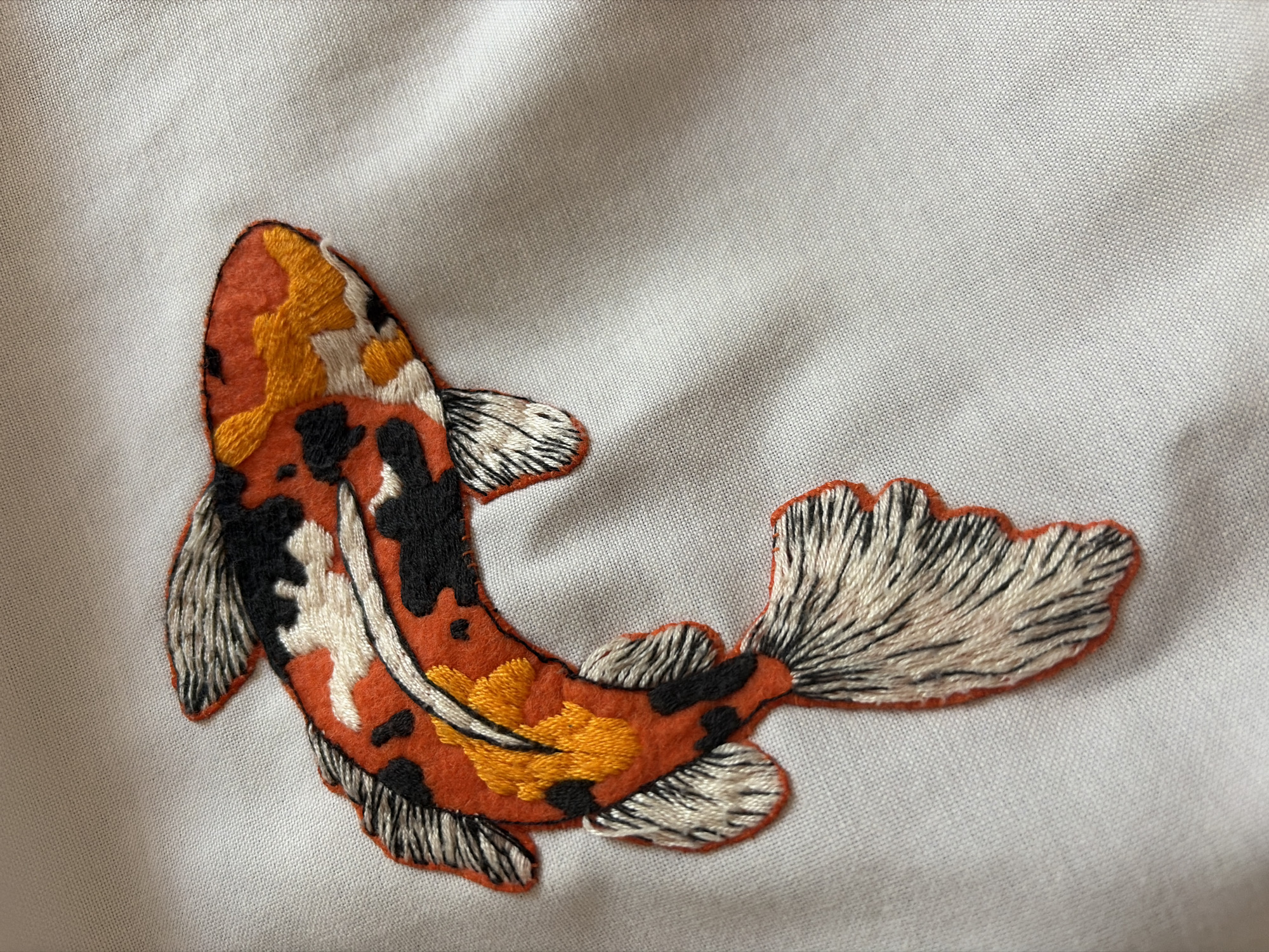 The image shows an embroidered koi fish on a light gray fabric. The fish is primarily orange with black, white, and yellow markings. The fins and tail are white with dark detailing to suggest texture and movement. No location information is available in the image.