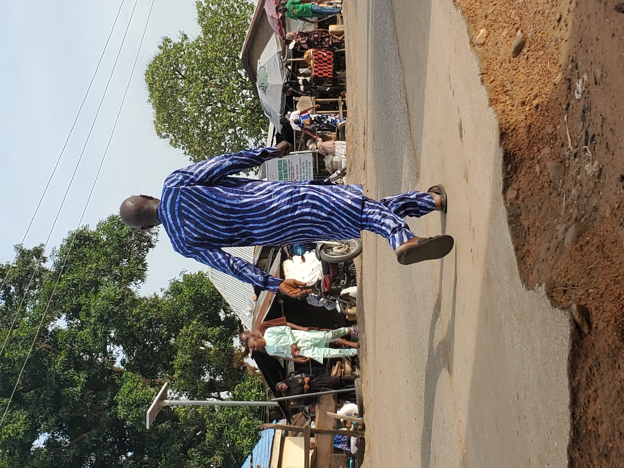 The image depicts a vibrant street scene in Mararaba, Nigeria, captured during daylight hours under a bright sky. The central figure is a man with a shaved head, dressed in a striking blue and white striped traditional outfit (likely an agbada) and sandals, seen walking along a paved road away from the camera, moving towards the right side of the frame.
To the left of the road, a bustling area features several makeshift stalls or shops with awnings, suggesting a market or commercial zone. Numerous people are visible in the background, including one individual in a light green traditional attire standing near a building, and others congregating around the stalls. A motorcycle is parked amidst these structures. The paved road gradually transitions into an unpaved, reddish-brown dirt area on the right.
Lush green trees with dense foliage frame the scene, appearing in the background and extending into parts of the foreground. Overhead utility lines are visible against the sky. A white rectangular sign near the stalls advertises medical services. The visible text on the sign reads: "AWARD MEDICAL C AND MATERNITY", "LONG BONA STREET" (or "BORNO STREET"), "Our Services:", "GENERAL MEDICAL SERVICES", "GENERAL LAB ANALYSIS", "ULTRA SOUND SCANNING", and "AND DENTAL CLINIC". The scene portrays everyday activity in a lively community setting.