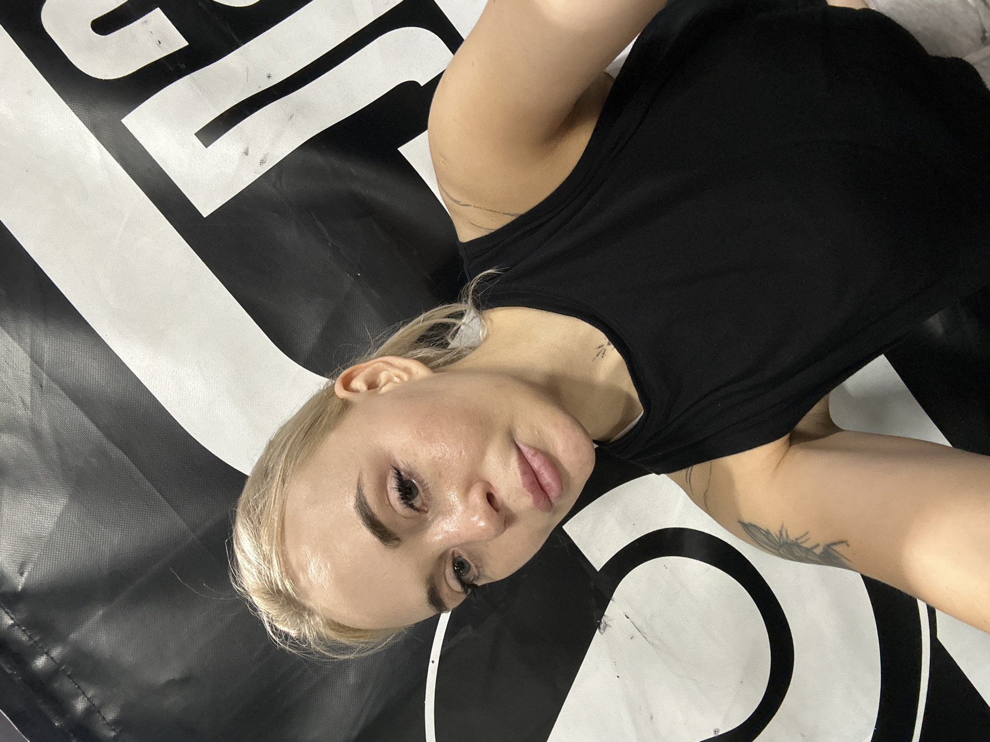 A woman with fair skin and blonde hair is lying down, seemingly indoors, on a black and white graphic surface. She is wearing a black tank top. The image is taken at a close range, focusing on her face and upper body. The graphic surface she is lying on consists of bold, abstract black and white shapes. There is a tattoo visible on her left upper arm and a smaller tattoo on her neck. The location is not discernable from the provided image, and there is no evidence to suggest it is in Megion, Russia.