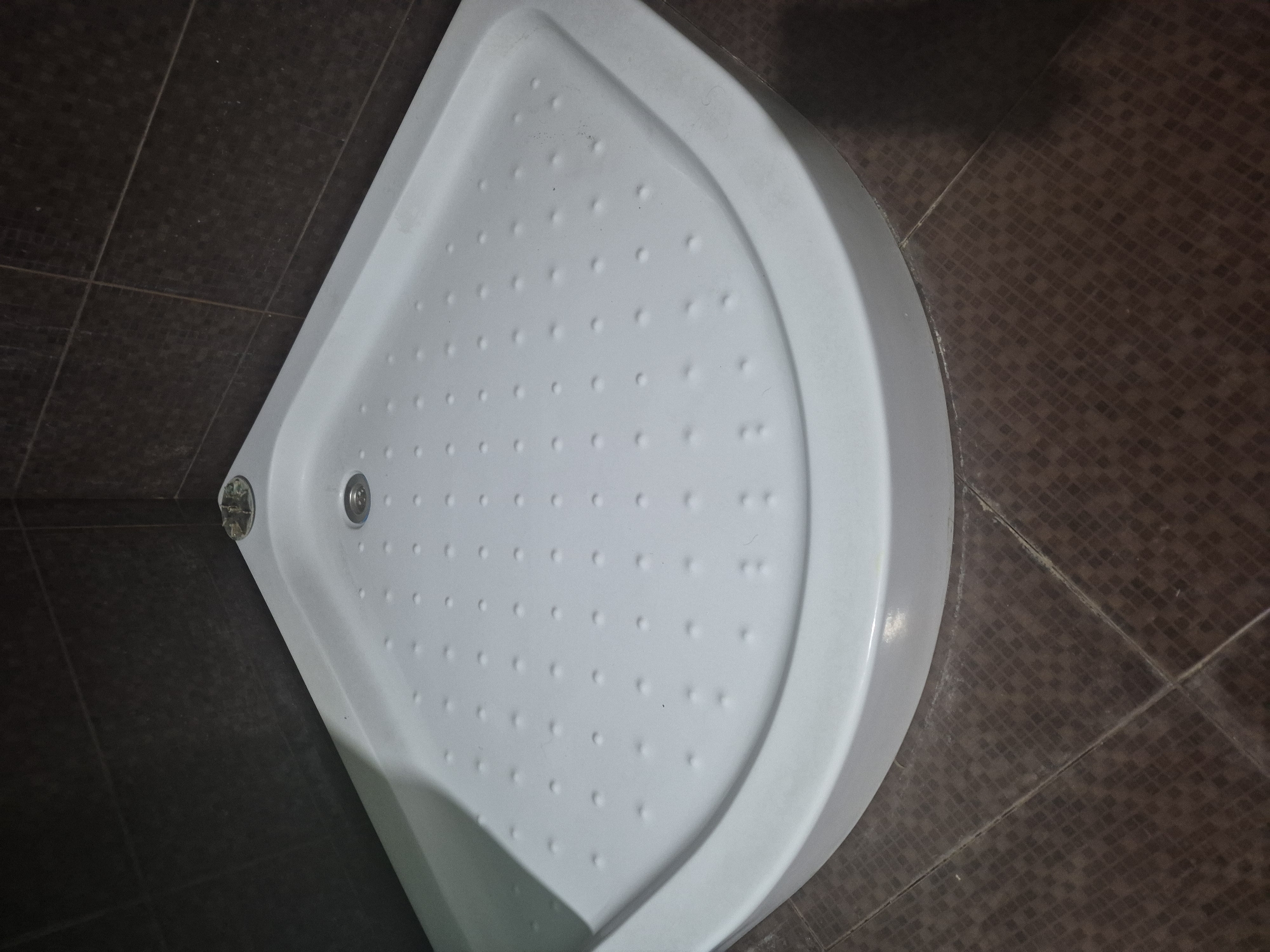 The image presents an overhead view of a white, quarter-round shower base or tray installed in a bathroom. The shower base features a textured, anti-slip surface with numerous small, raised circular bumps uniformly distributed across its floor. A circular metal drain is visible in the corner of the base, where the two straight edges meet.
The shower base is surrounded by dark brown floor tiles. These tiles, likely ceramic or porcelain, exhibit a subtle, slightly darker grid or small square pattern embedded within their surface. Lighter-colored grout lines delineate the individual tiles, creating a distinct pattern on the floor. Where the shower base meets the tiled floor, there is visible sealant or caulk, showing minor signs of discoloration or wear.
The scene is static, with no activity or individuals present. The lighting suggests an indoor setting, likely artificial. There is no visible text in the image. The setting is a bathroom in Yerevan, Armenia.