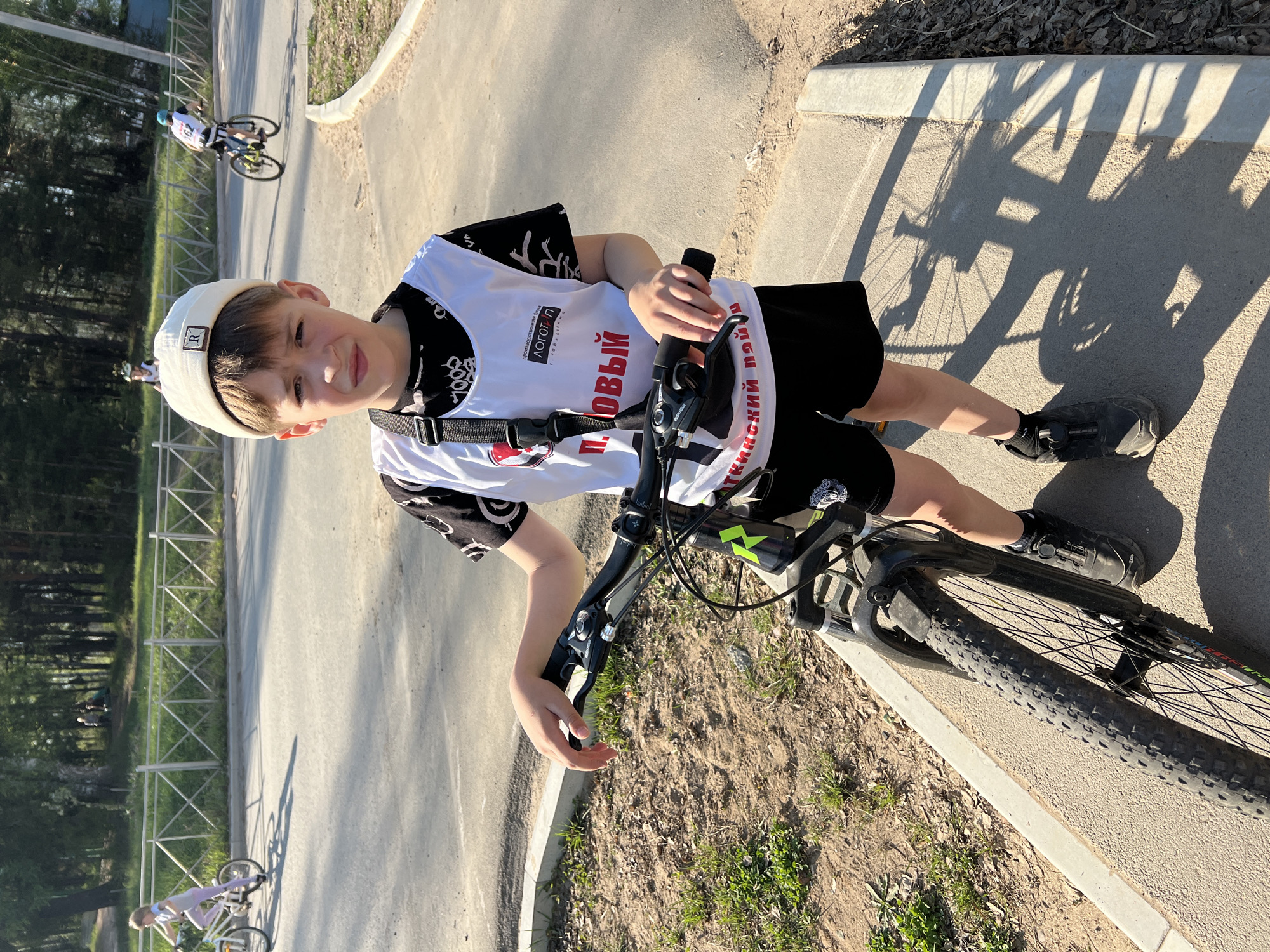 A young boy in a white cap and athletic jersey is standing on a black mountain bike on a paved path in Noviy, Russia. The jersey has the word "НОВЫЙ" in red Cyrillic script. He is wearing black shorts and black athletic shoes. Another cyclist is visible in the background on the same path. A metal fence runs along the edge of the path. The sun casts a long shadow of the bicycle on the path.