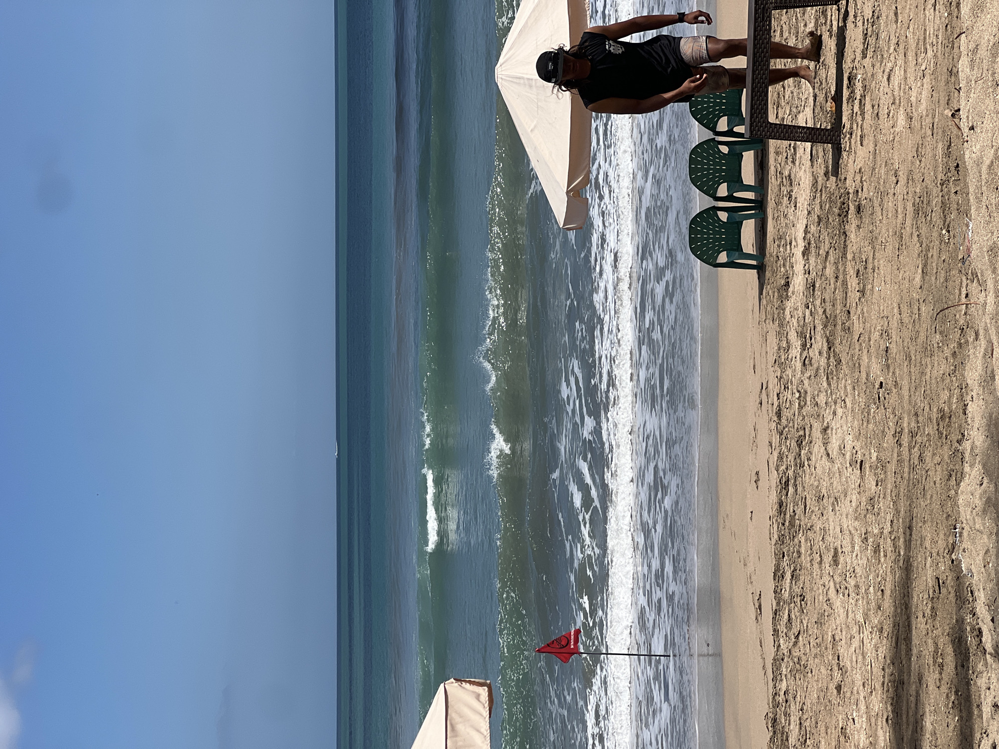 A wide shot shows a beach scene in Kuta, Indonesia, under a clear blue sky. The ocean is a deep turquoise with white-capped waves breaking on the shore. In the foreground, the sandy beach is littered with dried leaves and debris. Three green plastic chairs sit on the sand near a low, brown table. A man in a black tank top and patterned shorts walks from right to left, partially obscured by a large, light-colored umbrella. To the left of the frame, another umbrella is visible, and a red flag with text on a pole stands in the shallow water. In the distance, a small white boat is visible on the horizon.