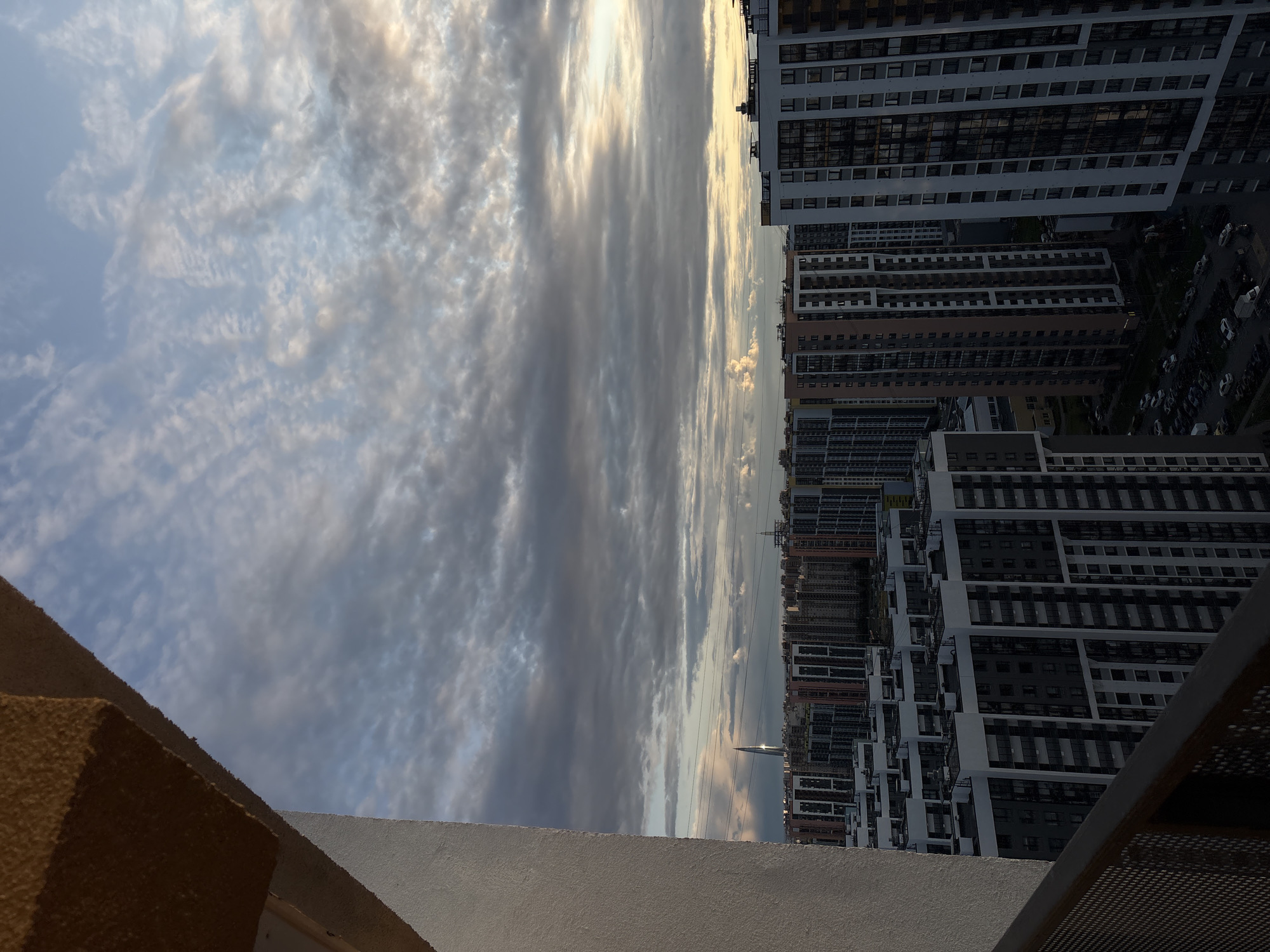 A high-angle, full shot captures a vast expanse of a city skyline under a cloudy sky. The foreground is dominated by the textured, beige underside of a building overhang and a section of a white wall, obscuring the bottom left portion of the image. This architectural element juts diagonally from the bottom left towards the top center.
Beyond this, the city unfolds with rows of tall, modern apartment buildings. These structures are predominantly white, grey, and some are accented with muted orange and brown tones. They are densely packed, creating a uniform urban landscape. Many of these buildings feature dark-tinted windows. Below the buildings, a street level view reveals a dark parking lot filled with numerous cars and a few vans.
In the distance, a slender, illuminated skyscraper pierces the horizon, catching the light. The sky above is a canvas of soft blue and grey clouds, with patches of lighter sky, suggesting either dawn or dusk. Subtle hues of yellow and orange are visible on the horizon, indicative of the setting or rising sun. Overhead power lines are faintly visible across the middle ground of the cityscape. The scene appears to be an urban residential area.