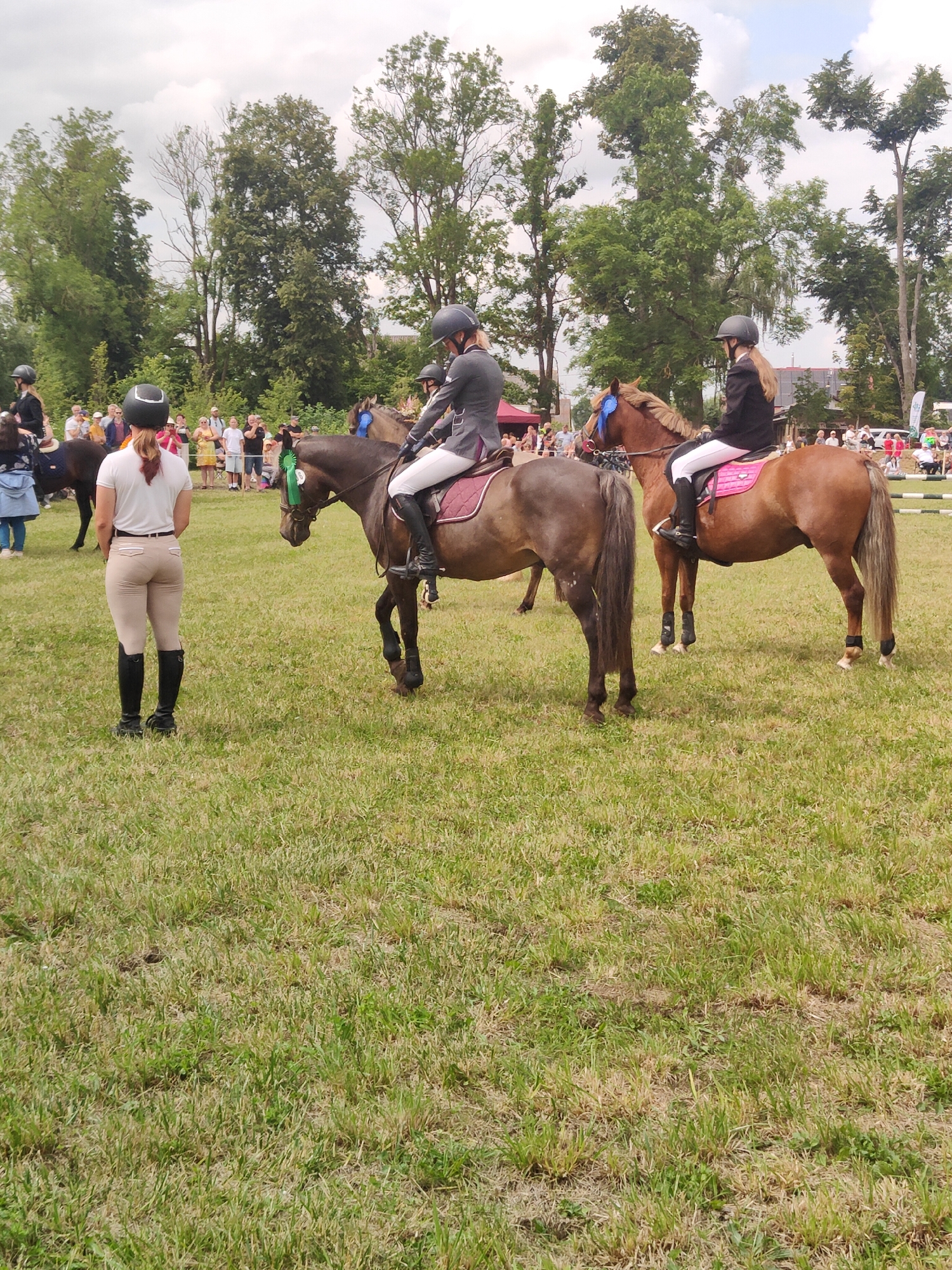 In Žagariškiai, Lithuania, an outdoor event featuring equestrian activities is underway on a grassy field. Several individuals are present, primarily young women dressed in riding attire, including helmets, riding jackets, and breeches. Two women are mounted on horses, one on a light brown horse in the foreground to the right, and another on a darker horse to the left. Both horses are adorned with blue ribbons, suggesting a competition or award ceremony. A third woman in similar riding attire stands on the grass in the foreground, facing away from the camera, her hands resting on her hips. In the background, a crowd of spectators gathers, observing the event. Trees line the perimeter of the field, and the sky is overcast. The environment is characteristic of a rural setting, possibly a park or a dedicated equestrian facility. The presence of people, horses, and a formal riding setup indicates a structured event, likely a horse show or competition.