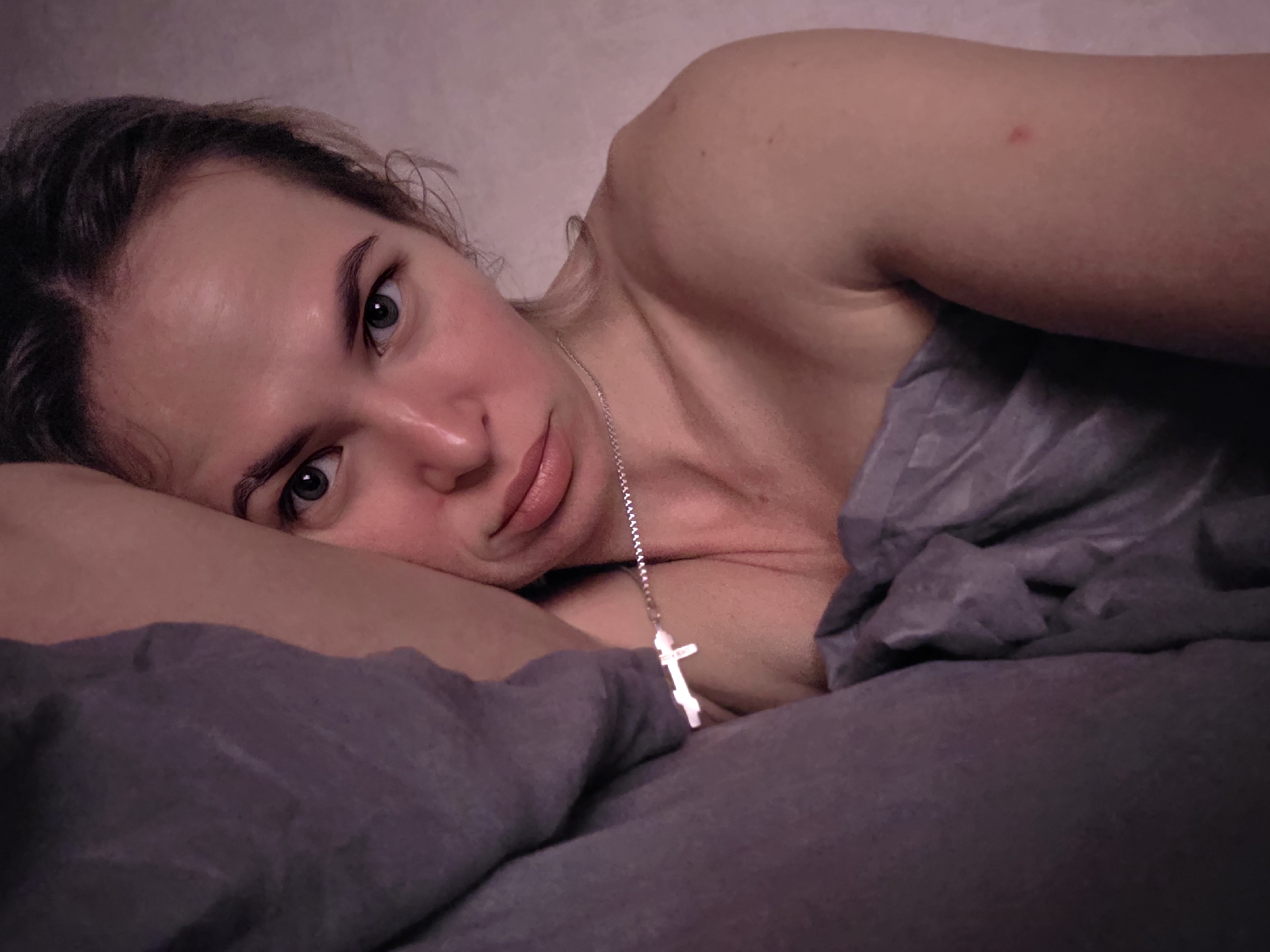 A female individual is depicted lying on her left side in a dimly lit indoor setting, likely a bed, in Chaikovsky, Russia. Her head is resting on a dark gray pillow or bundled bedding. She has light skin, dark hair, and light-colored eyes, which are open and directed forward towards the viewer. A silver-colored chain necklace with a small, plain silver cross pendant hangs around her neck. Her upper body is partially exposed above dark gray bedding that covers her lower body and part of her right side. A small, singular red mark is visible on her right upper arm/shoulder. The background consists of a light, textured wall.
