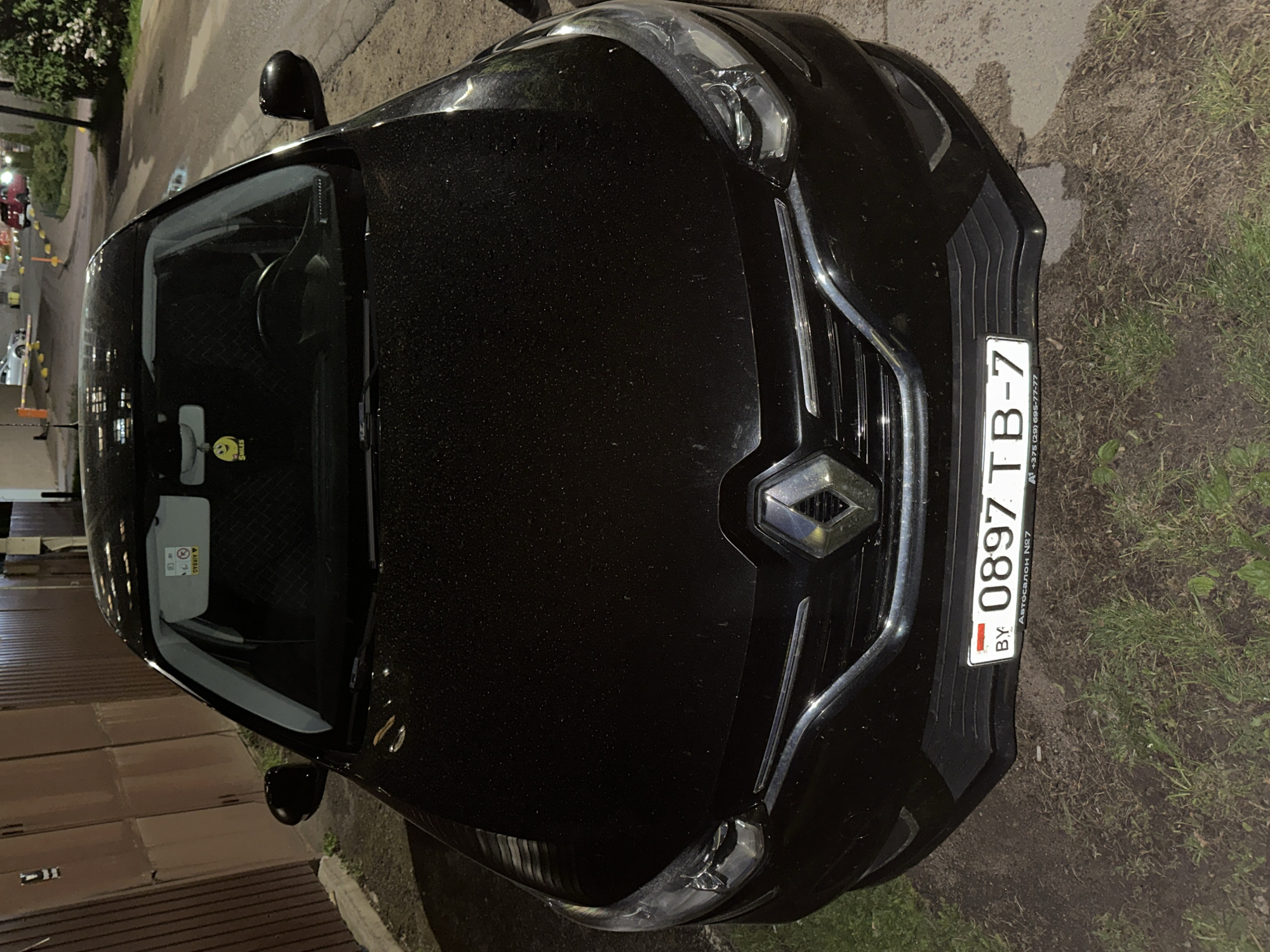 Here is a detailed description of the image:
The image is an overhead shot of the front of a black Renault car, likely taken at night, showing the hood, windshield, and front grille. The car has a license plate that reads "0897 TB-7" with "BY" indicating it's from Belarus. The plate also has "Автосалон №7" and a phone number "+375 (29) 695-77-77" written on it, suggesting it might be related to car sales from dealership number seven. The car is parked near a curb and some vegetation, potentially grass or weeds. There's a side mirror visible on the left side of the frame. The car has a smiley face on the windshield. In the background, there is a street with buildings and some traffic cones, suggesting an urban setting. Considering the Belarusian license plate, the location is likely in the city of Minsk, Belarus.