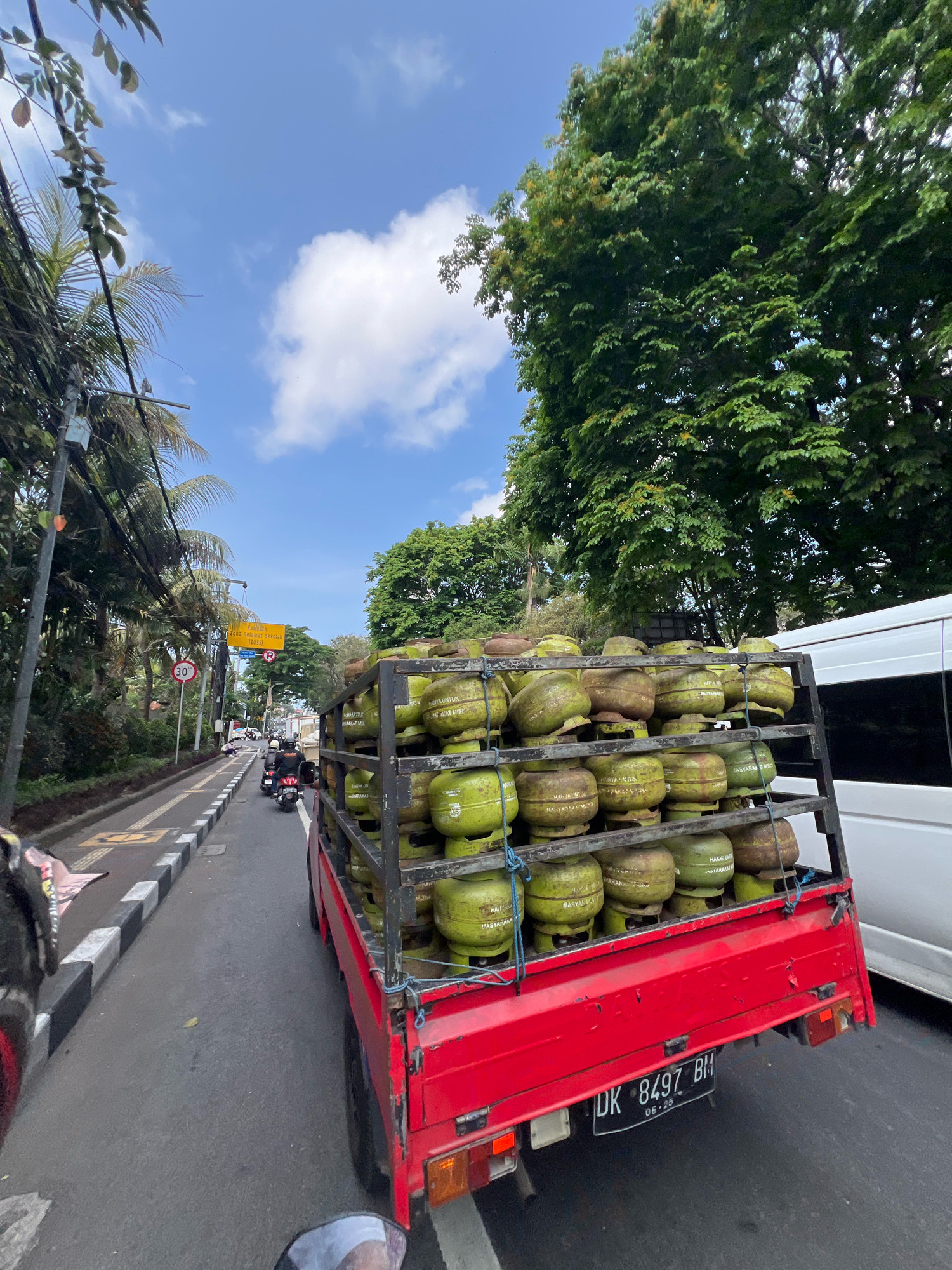 The image depicts a street scene in Denpasar, Indonesia, on a sunny day. A red truck, filled with a large number of green propane gas cylinders, is the central subject, driving on the right side of a paved road. The truck's license plate reads "DK 8497 BM". To the left of the truck, a lane is marked with a white and black striped curb, separating it from a sidewalk and vegetation. Motorbikes are visible in the traffic, suggesting movement. Overhead, a yellow sign with Indonesian text and a red circle with a line through it (indicating a speed limit of 30 km/h) is visible. Lush green trees line the street, and a bright blue sky with scattered clouds forms the backdrop. A white van is partially visible to the right of the truck.