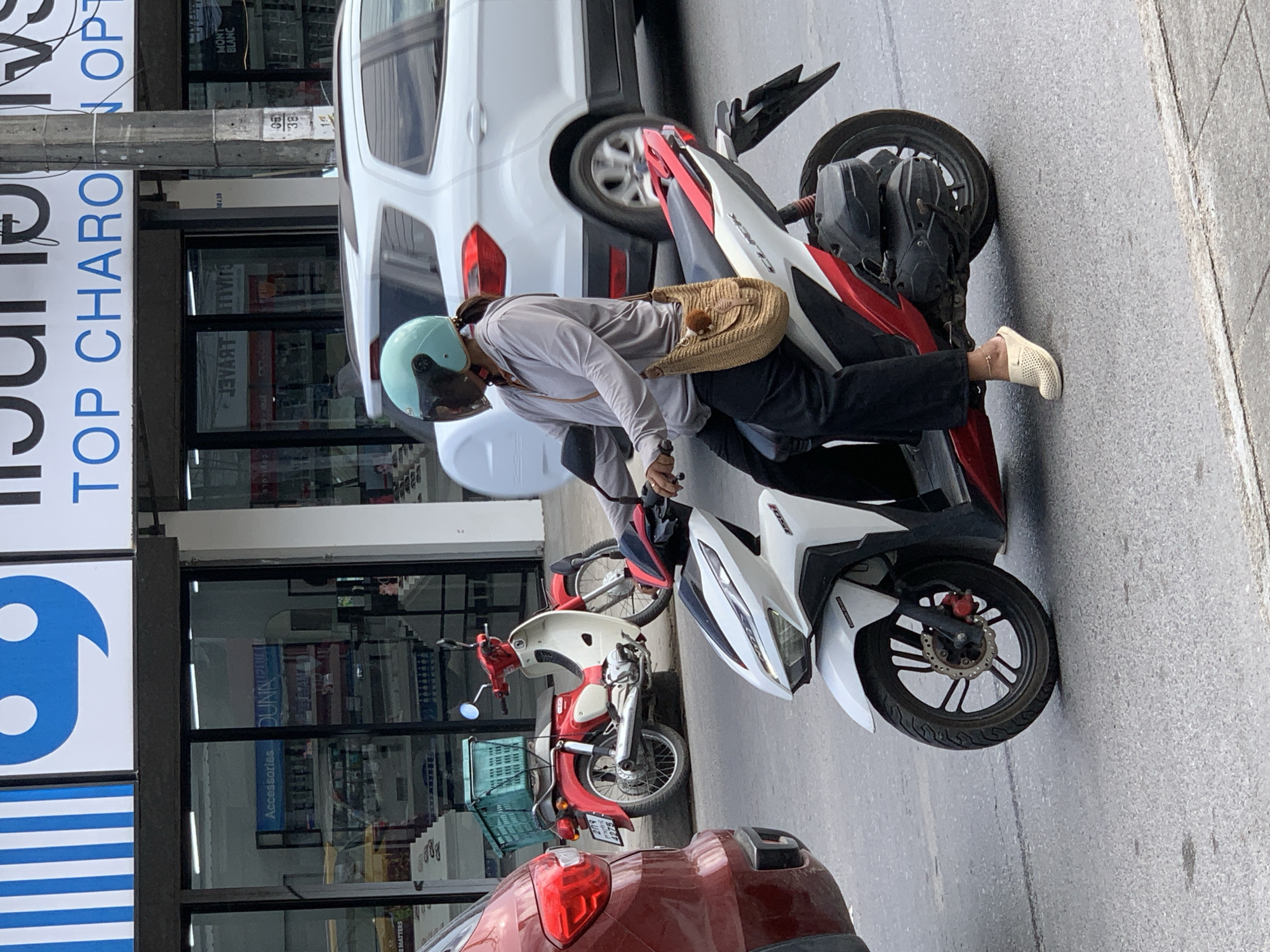 The image, taken in the city of Ko Samui District, Thailand, shows a woman riding a white and red scooter. She is wearing a light gray long-sleeved shirt, black pants, and light-colored slip-on shoes. On her head, she wears a light blue helmet. A woven straw bag with a fuzzy brown pom-pom charm hangs from her right shoulder. The scooter has a black engine and exhaust pipe, and a black rear fender. In the background, a white car is visible, as is a red car to the left of the frame. A red and white scooter with a green basket is parked behind the woman's scooter. The scene appears to be on a street with buildings lining the background, featuring signs with Thai text and some English text, including "Accessories" and "TOP CHARO." The road surface is asphalt.