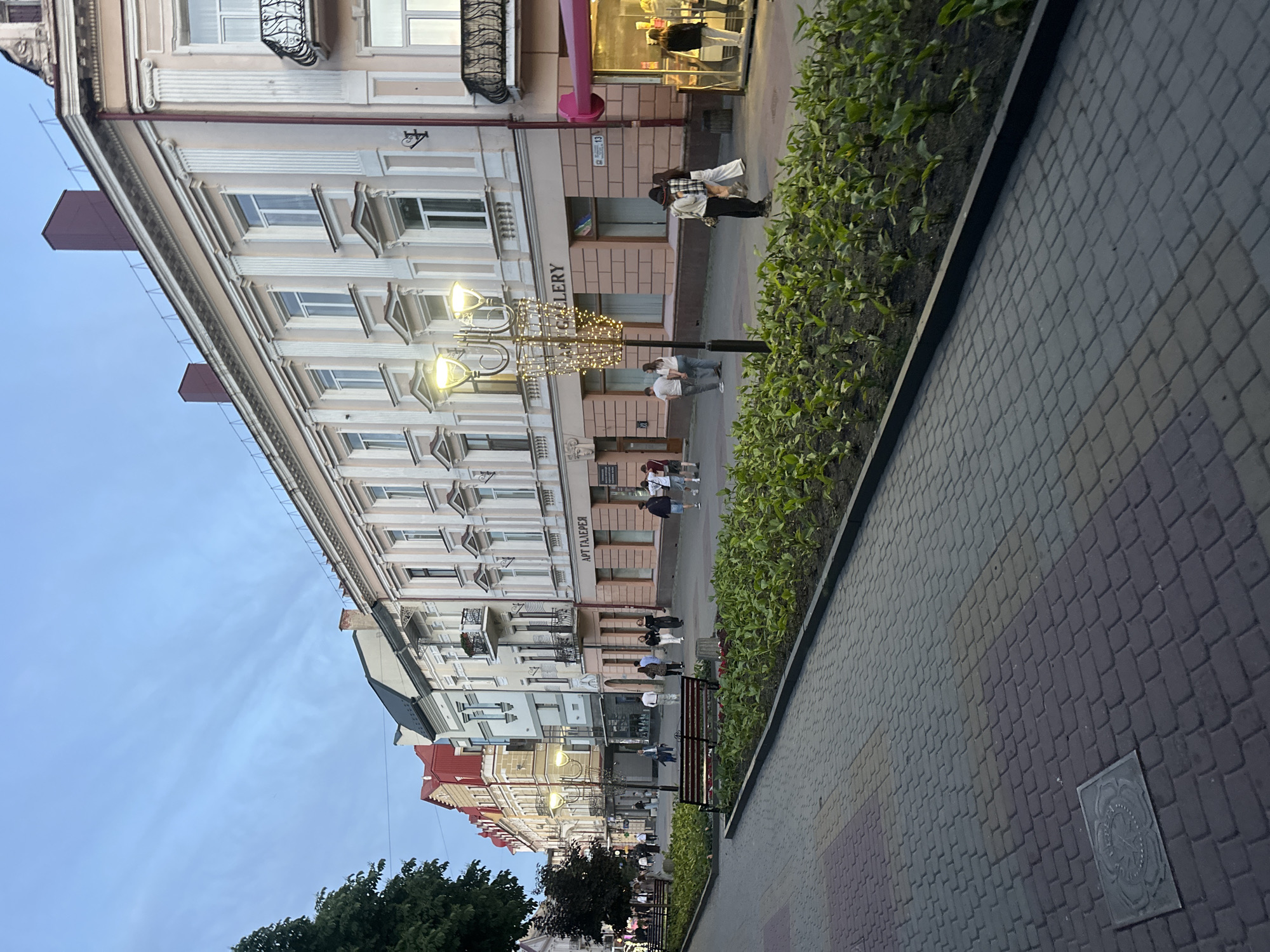 The image captures a pedestrian street in Ternopil, Ukraine, at dusk or early evening. The sky overhead is a muted blue with faint, wispy cloud streaks.
The street features a paved walkway constructed from interlocking pavers in varying shades of dark grey, light grey, and reddish-brown. A square, dark metal manhole cover with an embossed circular design is embedded in the paving on the left side. Long, rectangular planter beds filled with small, dense green foliage line both sides of the walkway. Several dark-framed wooden benches are positioned along the left planter.
Buildings line both sides of the street. On the right, structures display light peach or pink facades with white classical architectural details, including pilasters, ornate window frames, and cornices. Black wrought-iron railings adorn a visible balcony. A ground-floor establishment is marked with "АРТ ГАЛЕРЕЯ" in Cyrillic script and "GALLERY" in Latin script. A small kiosk with a vibrant pink canopy is partially visible on the far right. Buildings on the left side exhibit diverse designs, including multi-story structures with red and dark grey gabled roofs.
Multiple streetlights are illuminated, casting a warm light. A prominent black streetlamp in the right foreground features multiple ornate arms and is elaborately decorated with a cage-like structure of numerous small, twinkling string lights.
Numerous individuals populate the street, walking in various directions. People are observed singly, in pairs, and in small groups. For instance, near the decorative streetlight, a group of three individuals (two women, one man), including a woman wearing a plaid shirt and a dark hat, are visible. Further down the street, additional pedestrians are seen strolling and interacting, indicating an active evening environment.