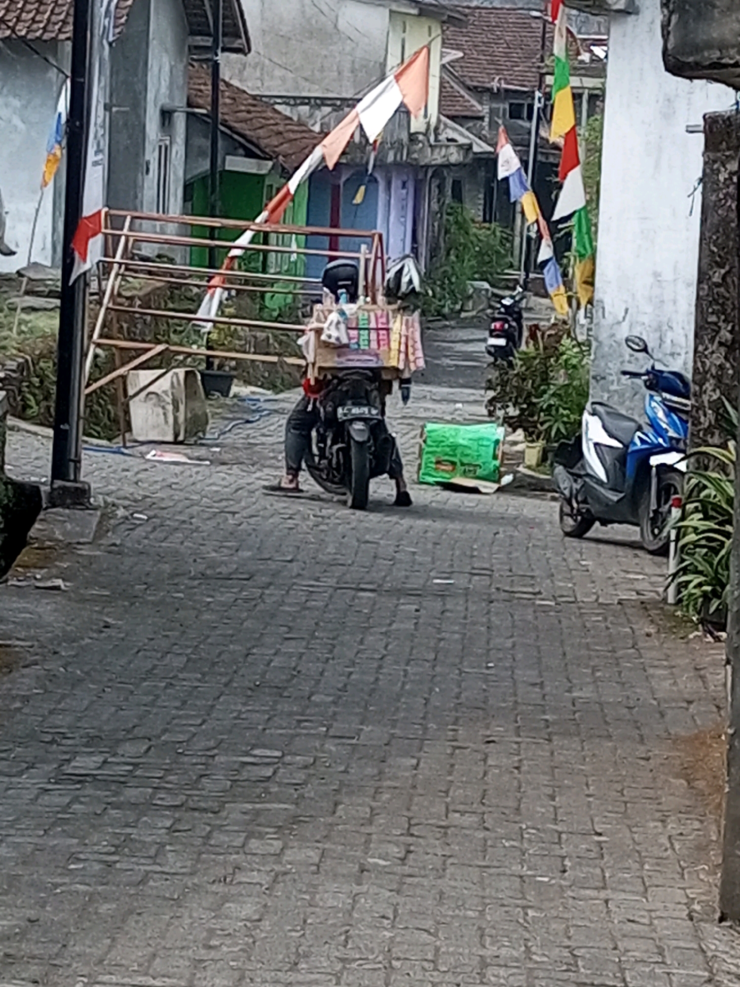 In Andongsili, Indonesia, a person wearing a helmet sits on a motorcycle loaded with items, parked on a cobblestone street. The motorcycle is positioned behind a metal barrier adorned with red and white flags. To the right of the motorcycle, a blue and white scooter is parked. In the background, buildings with tiled roofs and various flags are visible. The scene appears to be in an outdoor, possibly residential or market, area.