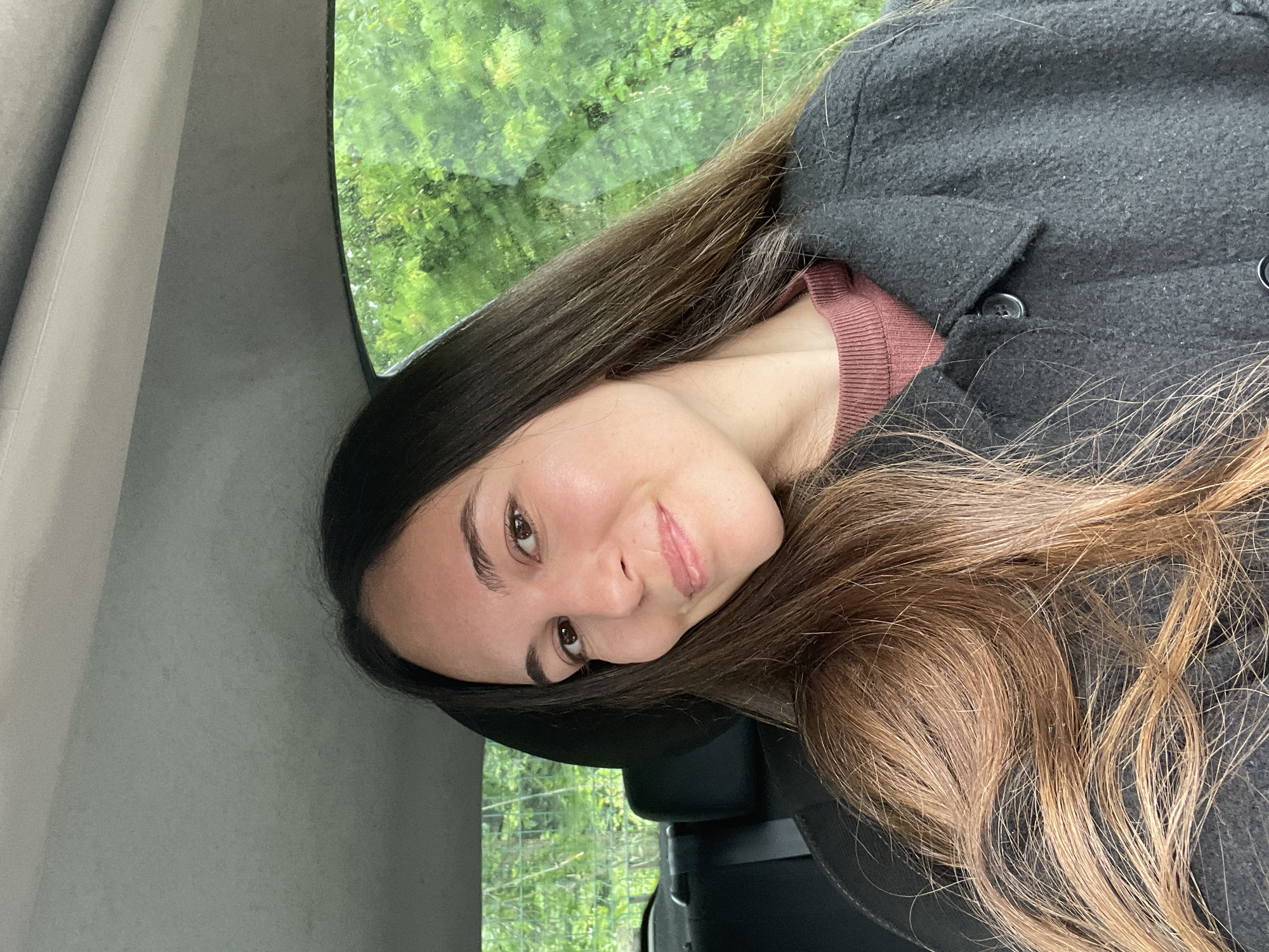 A young woman with long brown hair sits inside a car, looking at the camera with a slight smile. She is wearing a dark grey coat over a maroon-colored top. Behind her, outside the car window, is a view of green trees. The interior of the car is predominantly grey and black. It is not possible to determine the specific location in Sofia, Bulgaria from this image alone.