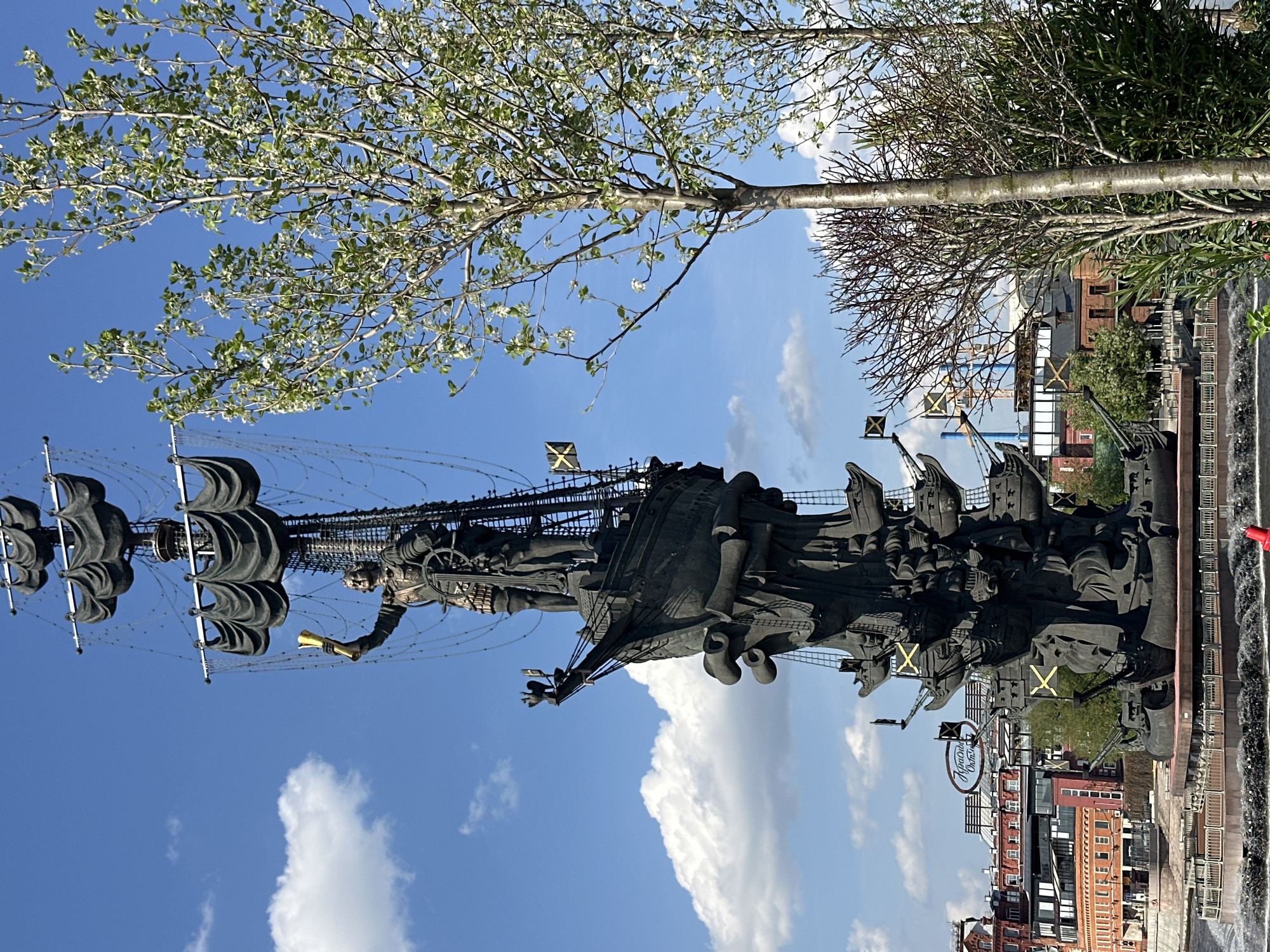 The image features a tall, dark-colored monument set against a blue sky with scattered clouds. The monument depicts a man standing on a ship, holding an object resembling a scroll or a trumpet. The ship has multiple masts and sails. Flags with a distinct "X" symbol are visible on the ship.
In the background, trees with foliage are present, along with buildings and structures that indicate an urban environment. A river or body of water is also visible in the lower part of the frame.
This statue is the Peter the Great Statue, located in Moscow, Russia.